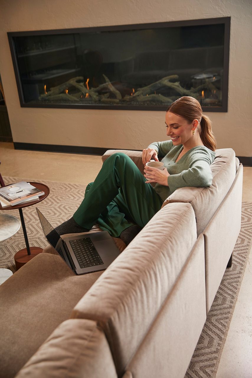 A smiling woman holding a mug sits on a sofa next to an open laptop, backed by large, wall-mounted fireplace. 