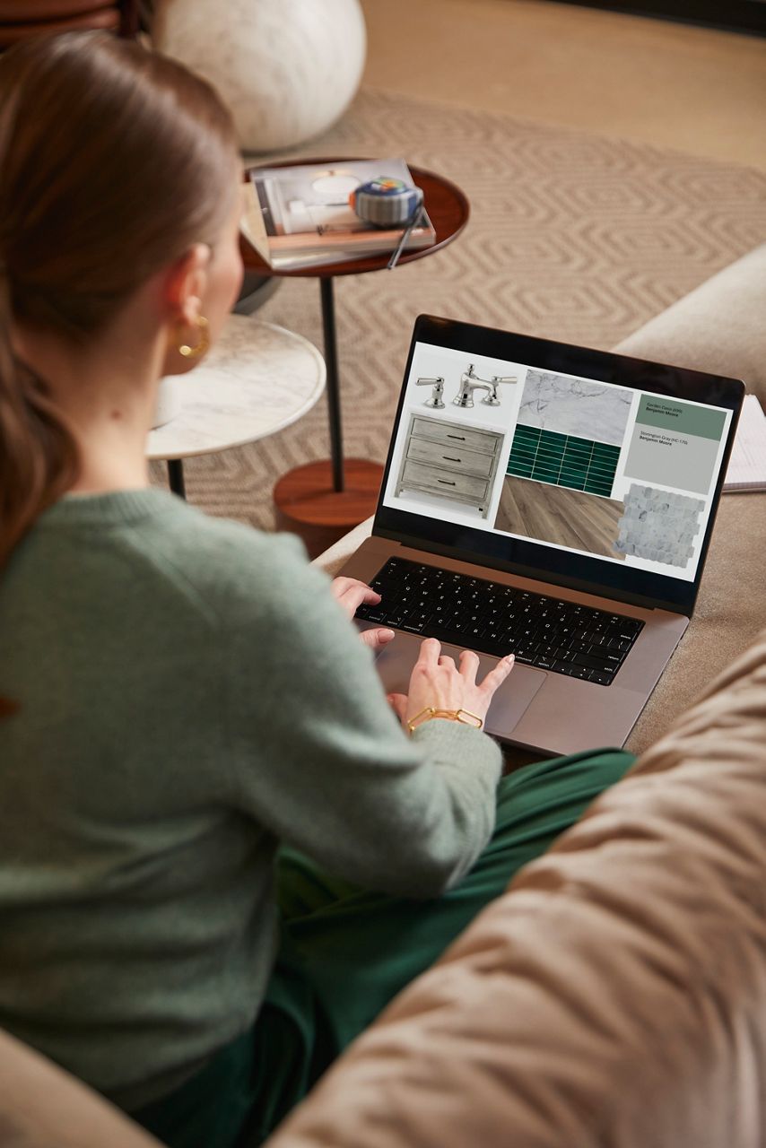 A woman sitting on a sofa and scrolling on a laptop with faucet, vanity, tile, countertop, color, and floor options on screen. 