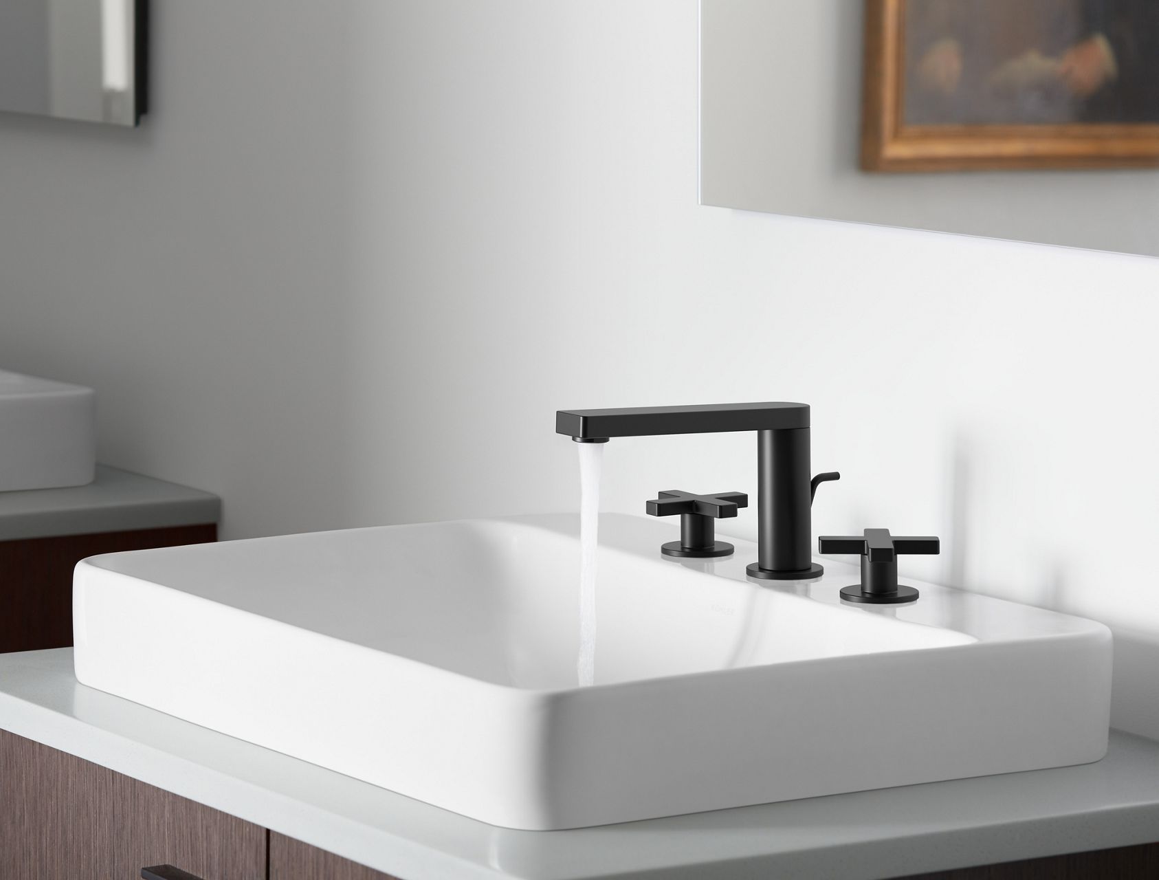 A matte black Composed widespread faucet pouring water into a white sink next to a white wall.
