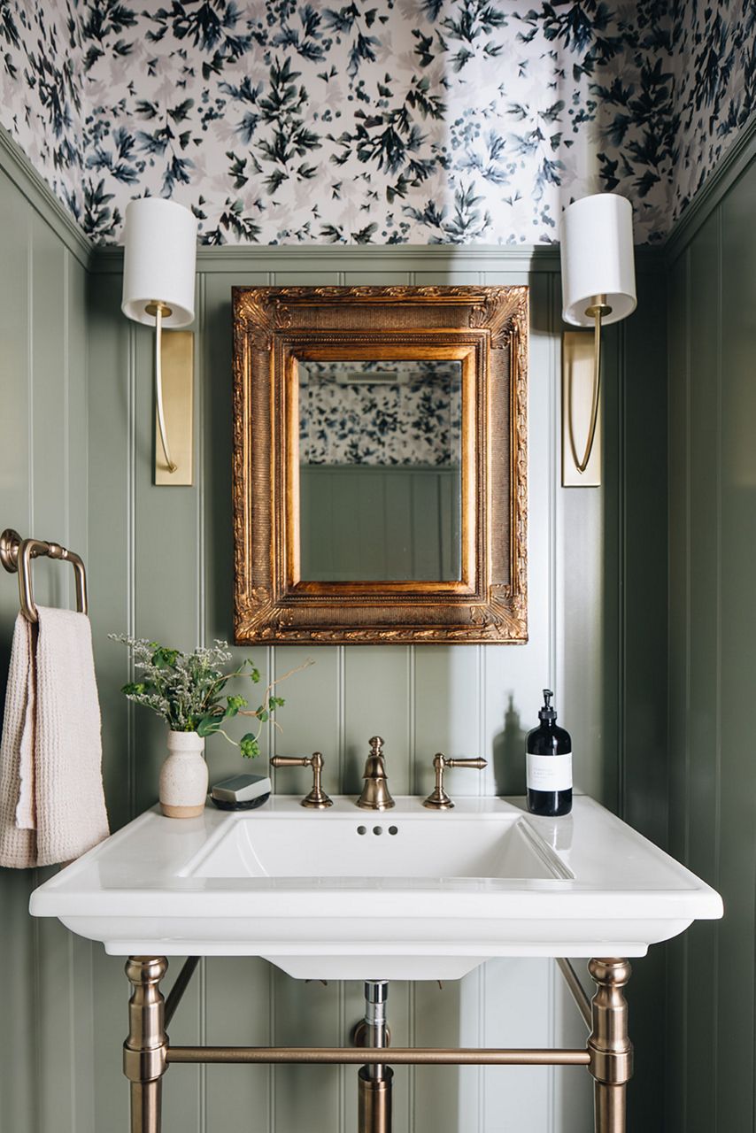 A bathroom with white KOHLER console sink on metal legs, sconces with white shades, a mirror in an ornate gold leaf picture frame, and sage green beadboard with blue and white floral wallpaper above it.
