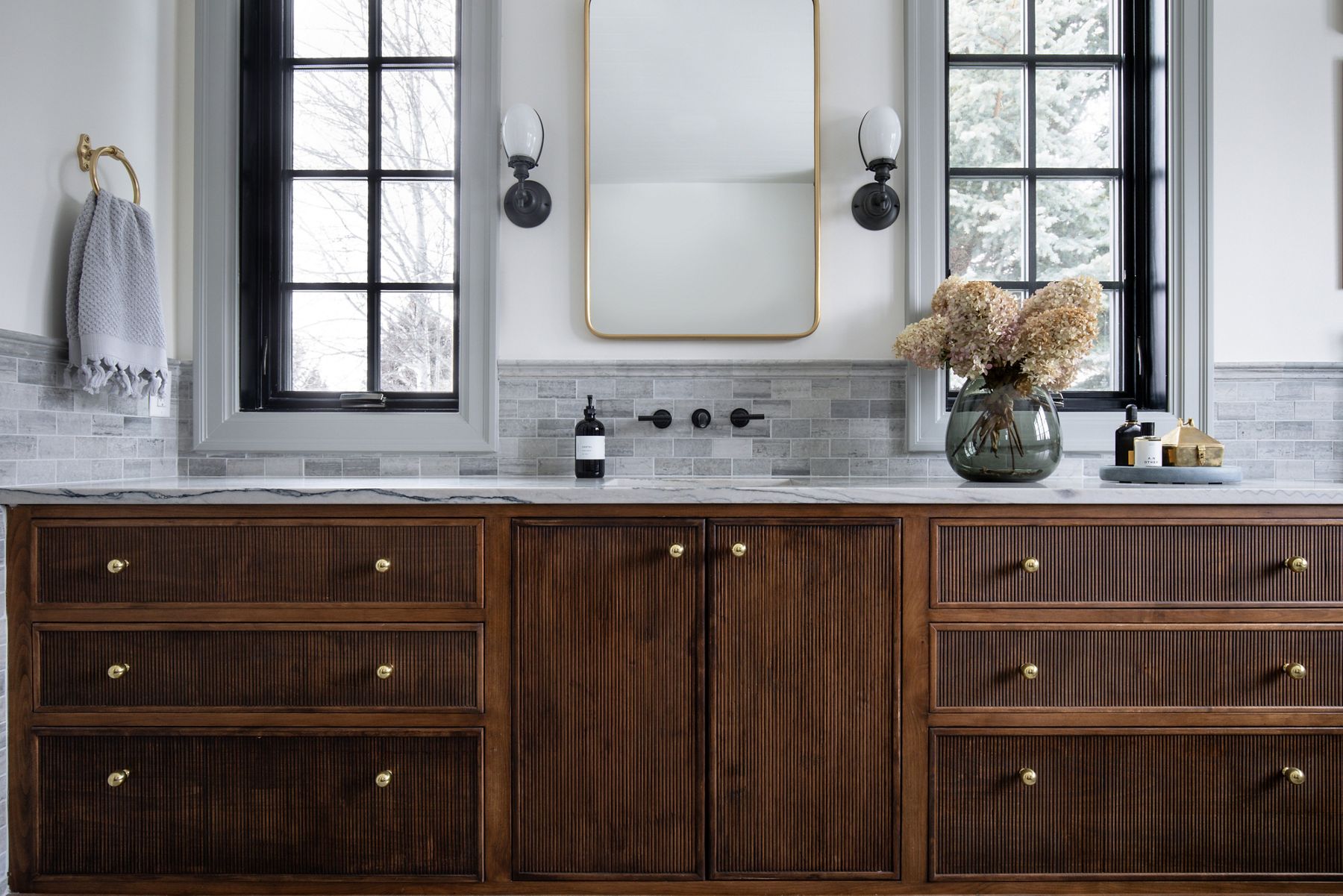 Windows and sconces with frosted glass shades bracket a gold-framed mirror above a KOHLER wall-mounted wide spread lever handle faucet mounted on  a marble tiled wall.