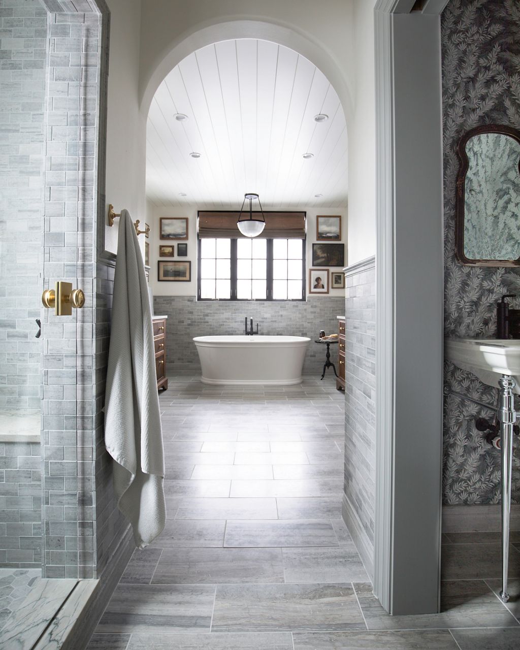 A white KOHLER freestanding bath stands under a window in a bathroom with classic elements like arched doorways, marble tile walk in shower, and neutral colors.