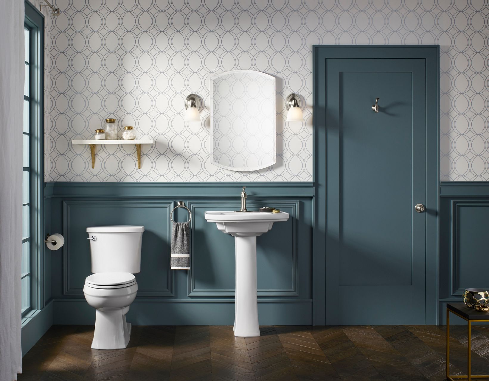 A KOHLER frameless medicine cabinet hangs in a newly designed bathroom alongside two single-light sconces, a wall-mounted shelf, a pedestal sink, and a KOHLER toilet.