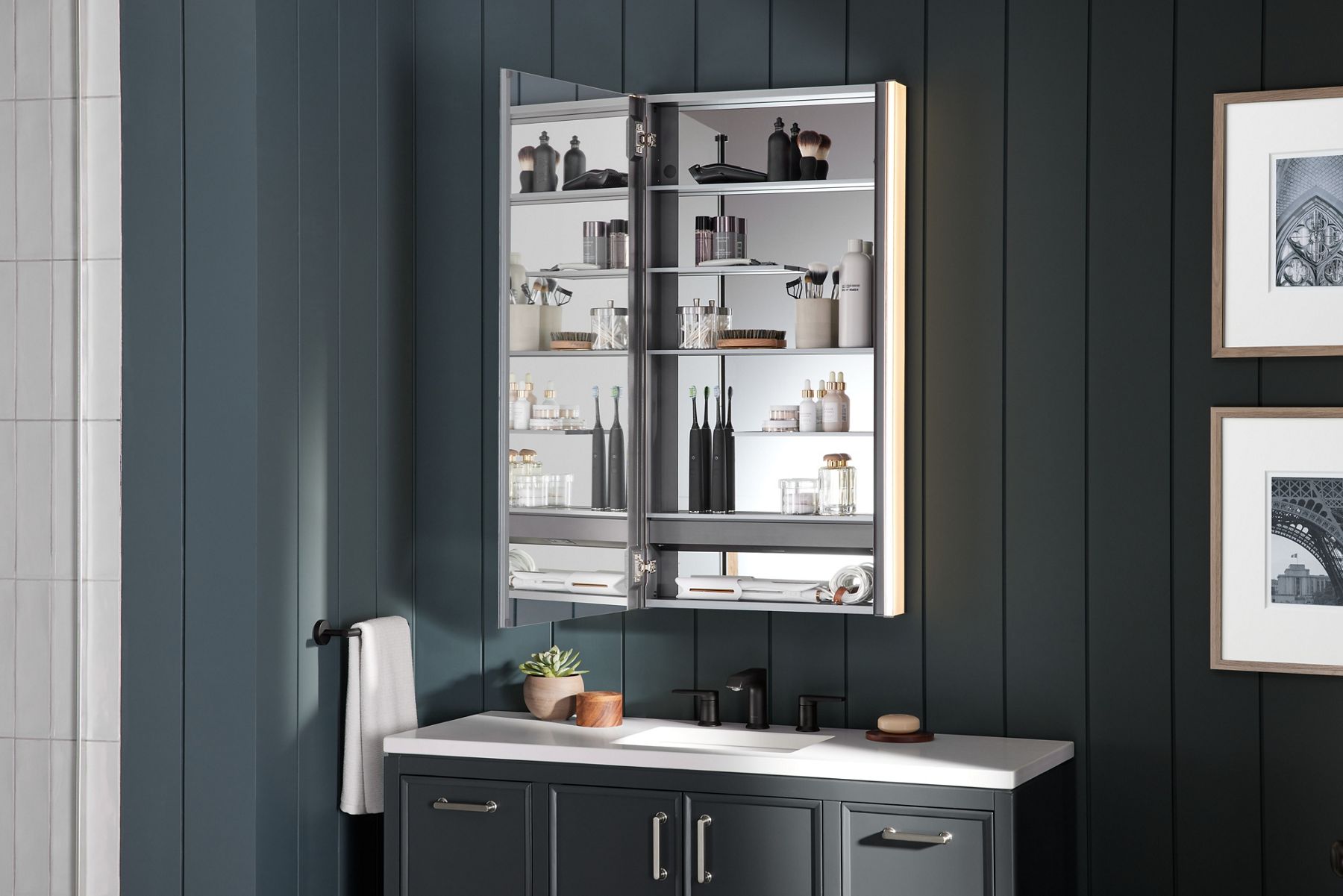 A medicine cabinet with its door open to reveal the mirrored interior surfaces and storage options, set against a dark gray paneled wall with a dark gray vanity with a  white counter top and black faucet.