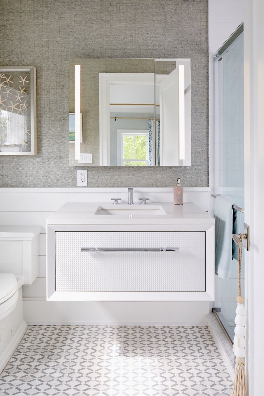 A white Enivo vanity with white top, polished chrome hardware and faucet and a lighted mirror in a bahtroom with patterned tile flooring, walls with lower ship lap and upper textured gray wallpaper.