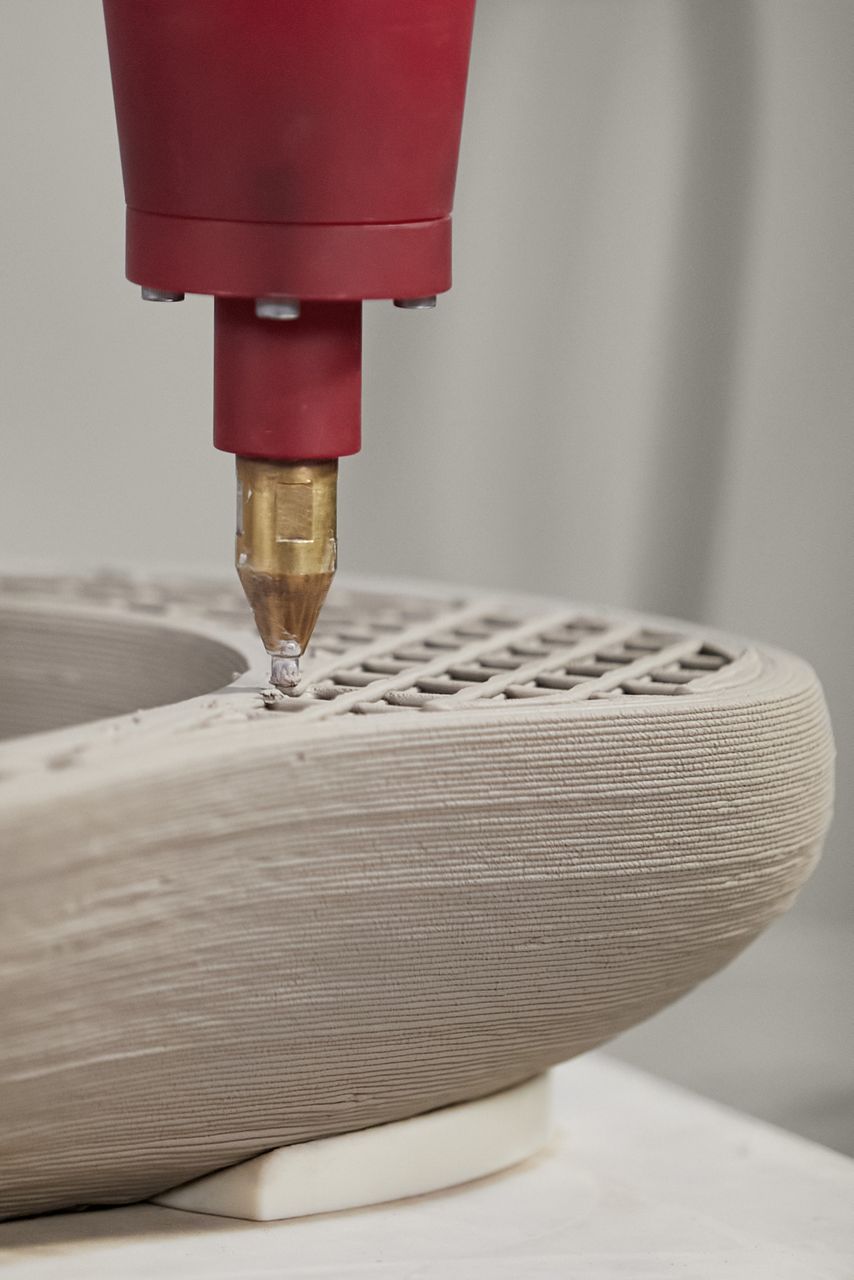 Close-up detail of a Rock.01 sink being 3D printed. The print nozzle is applying the vitreous china material to a partially completed sink, showing the internal bracing structure and the exterior layers from the printing process.