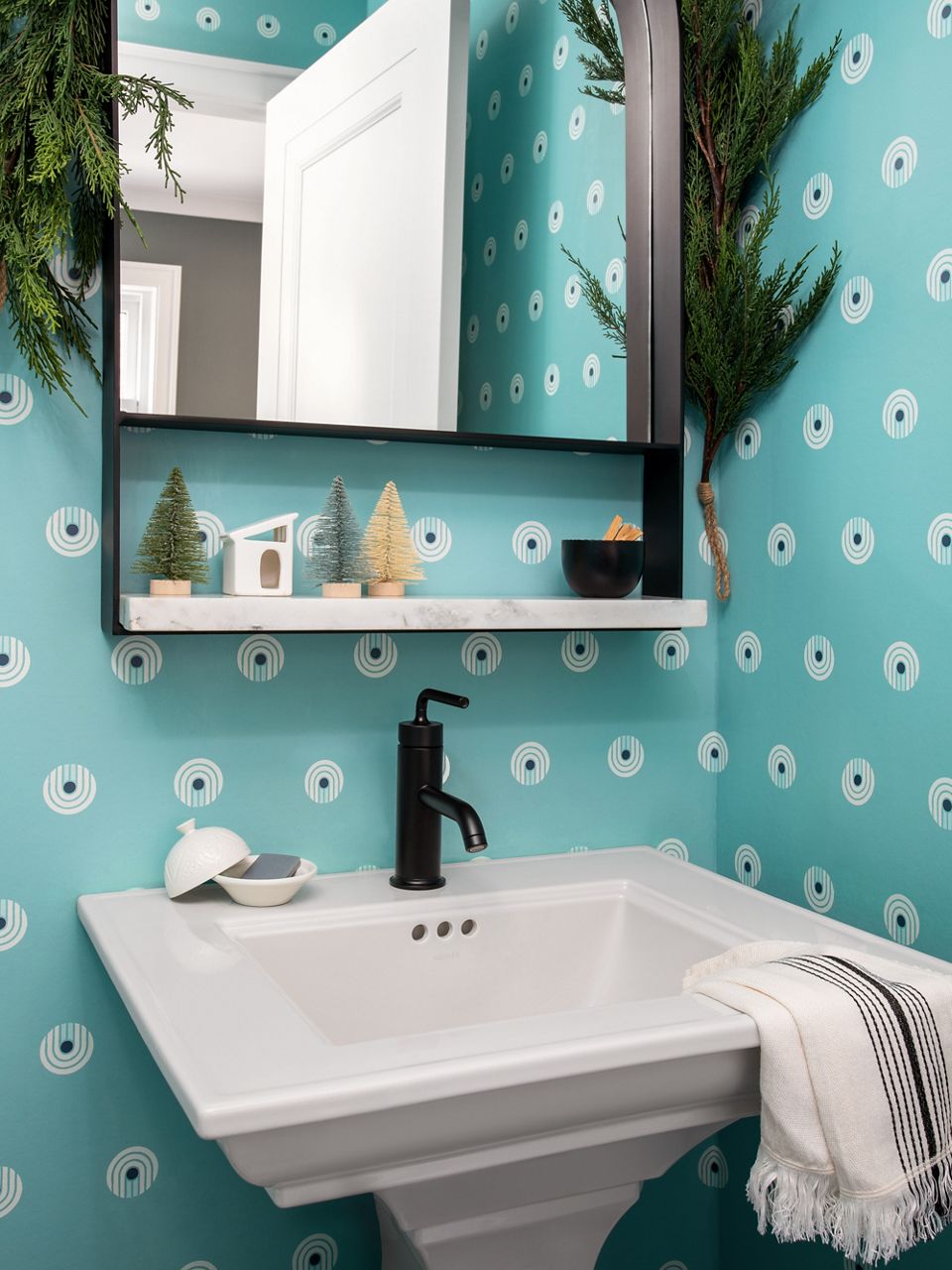 Image of a small powder room featuring a fun and funky, polka-dotted teal wall paper and white pedestal sink with matte black, single-lever faucet. Holiday greenery is draped on the mirror.