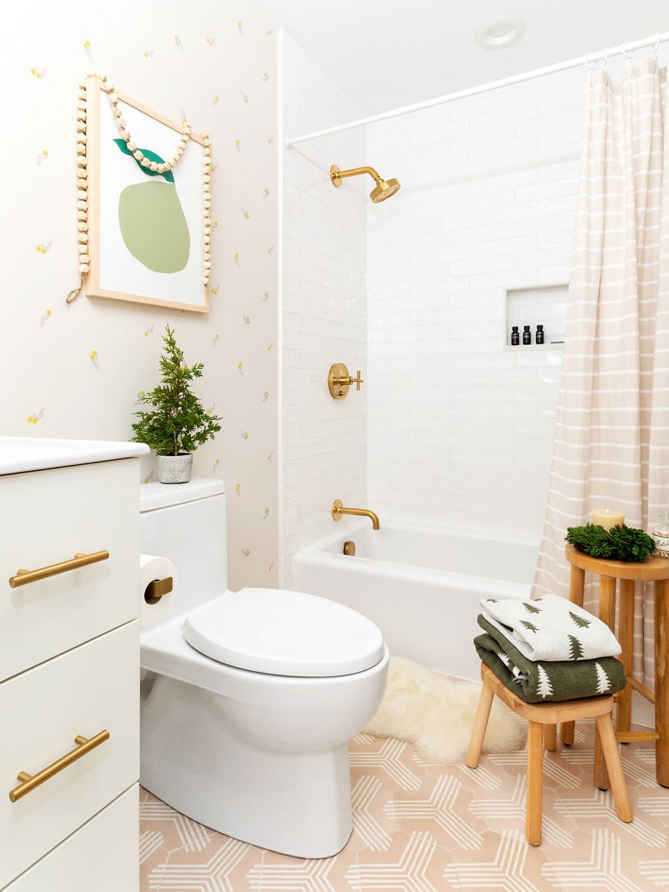 Wide image of a guest bathroom with white shower, one-piece toilet, white vanity and pops of brass on the shower showerhead, shower handles, and drawer pulls. Holiday greenery and Christmas tree-patterned towels create a festive environment.