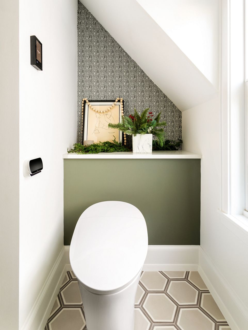 Image of a private toileting space is shown, featuring an intelligent toilet and wall-mounted remote control. Holiday greenery sits on the shelf behind the toilet.