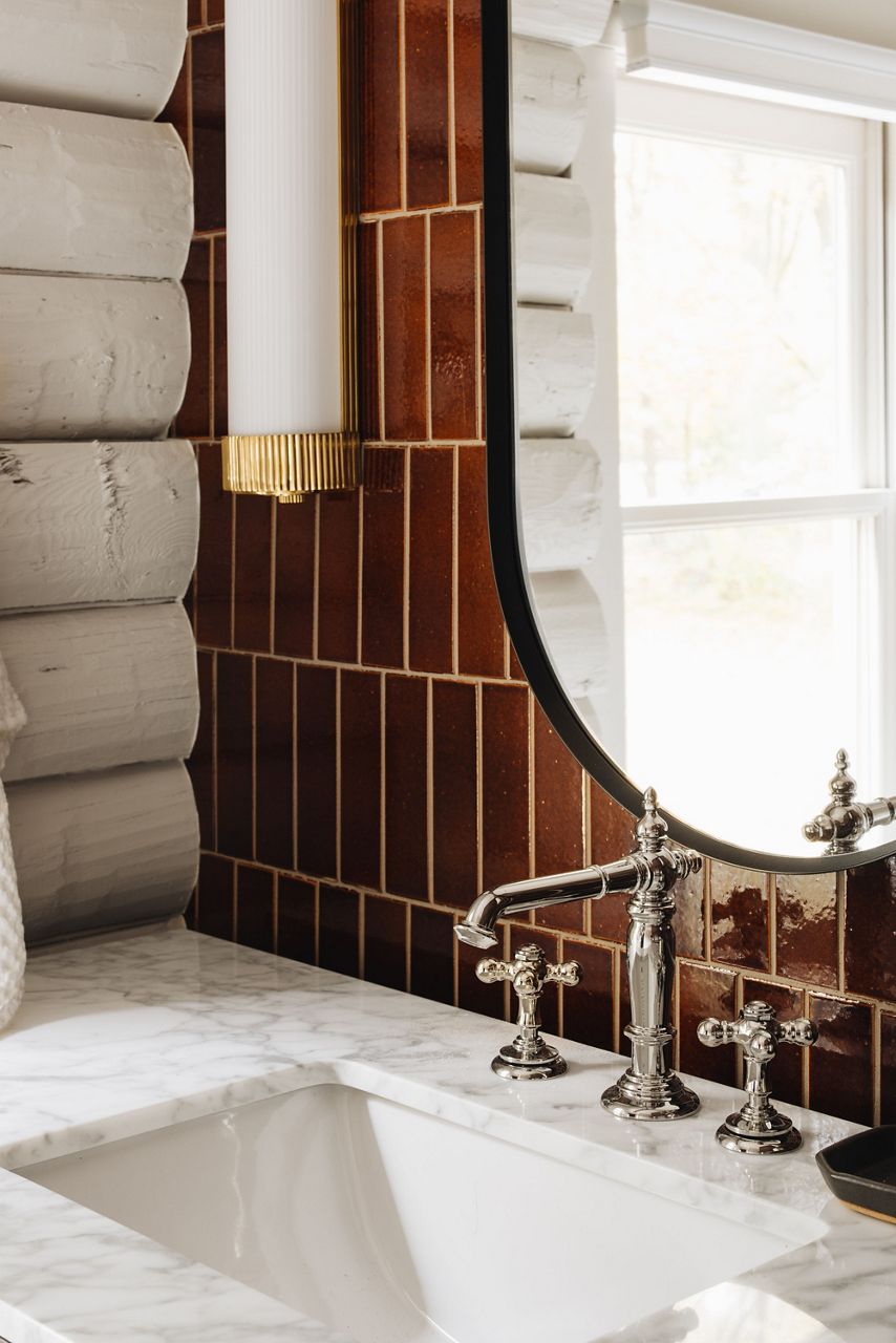 Image of the vanity area in the bathroom of the cabin, focused on the faucet and sink space to feature the Artifcats faucet and cross handles in polished nickel finish, mixed with matte black finish trim on the oval mirror and a brass accent finish on the lighting sconces.