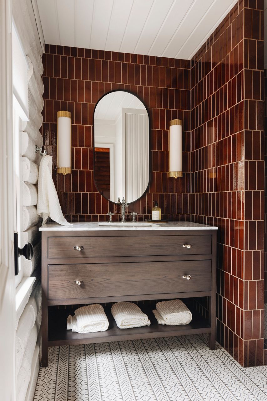 Image of the vanity space in the cabin bathroom where a rustic wood freestanding vanity with open towel shell on the bottom sits in front of a bold rootbeer-colored glossy tile wall from floor to ceiling. A Kohler Artifacts bathroom faucet in polished nickel finish is atop the white counter top, with vertical sconses on either side of the oval matte black rimmed mirror.