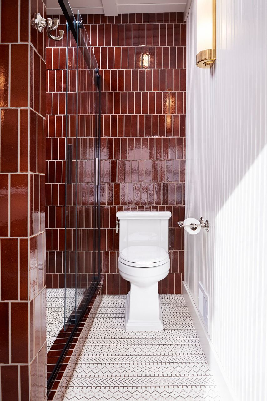 Image of the white Tresham toilet centered in front of the vertically tiled, rootbeer-colored wall and intricate geometric patterned white and black floor tile. The toilet paper holder on the wall is of polished nickel finish.