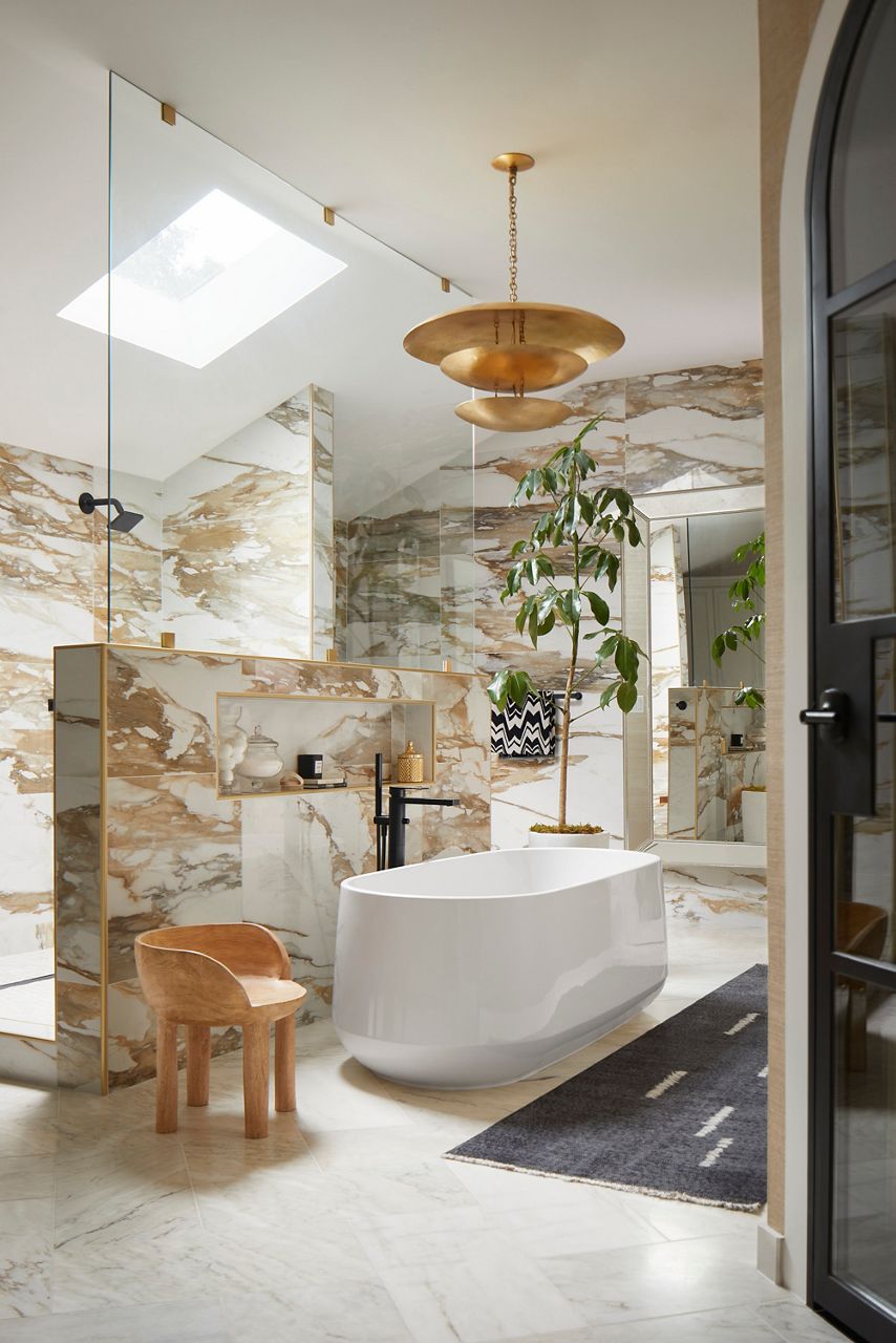 Image of Nikki Chu's primary bath suite featuring a white, oval Kohler freestanding bath as the centerpiece of the room. Above is a skylight in the vaulted ceiling and a gold chandelier. The tile walls have a gold and white marble tone, enclosing the shower area.