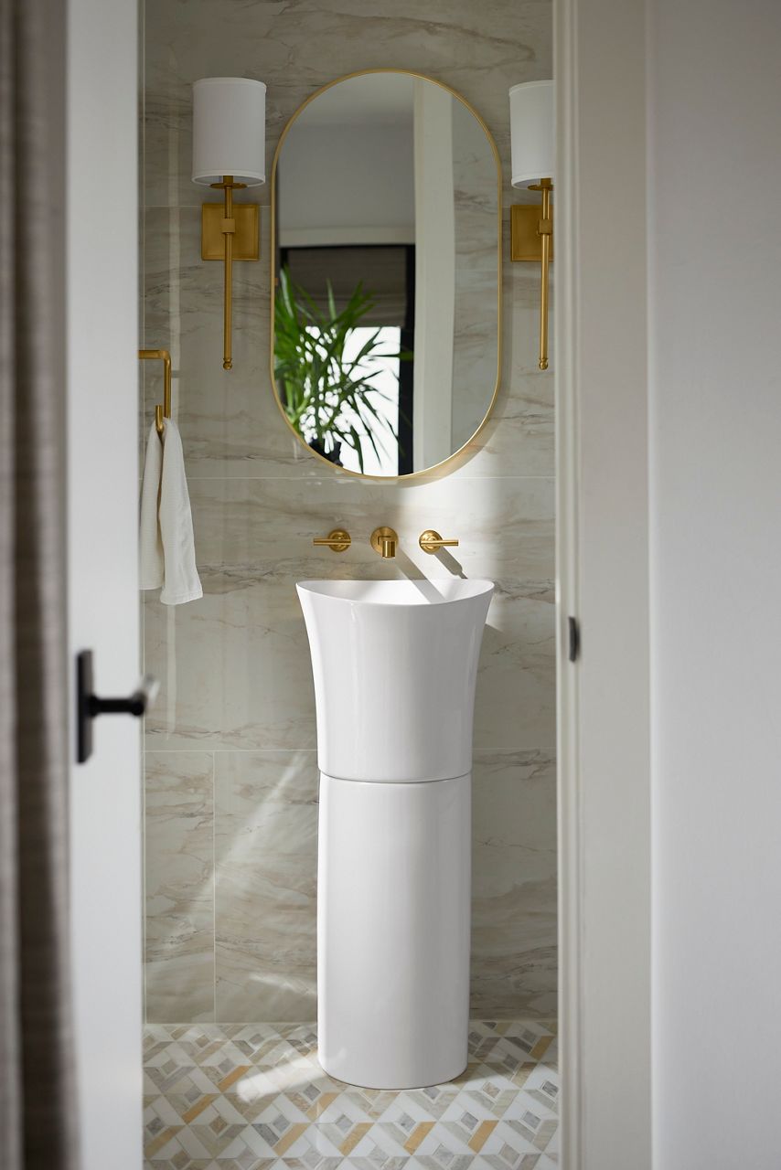 Image of Nikki's guest bathroom, centered on the wall is a vanity set up featuring the tall, white, oval pedestal sink from Kohler and a Kohler wall-mounted brass faucet. Above the sink and faucet are two matching brass sconces with white shades and an oval mirror with matching brass trim.