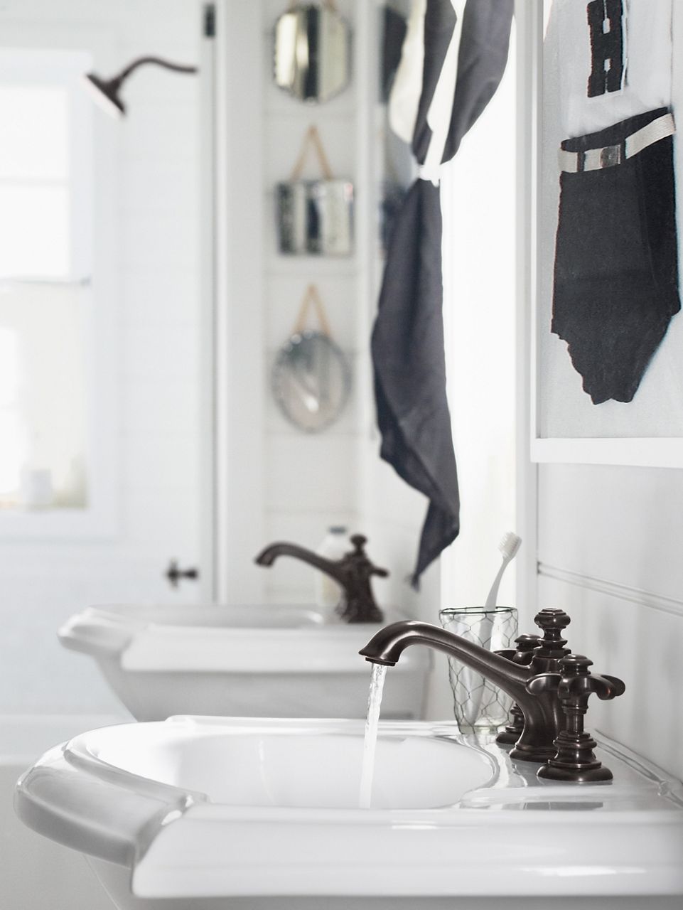 Water flows from a black bell spout KOHLER faucet with separate prong handles, set in a white pedestal sink with curved front, in a bathroom with white tile and black accents.