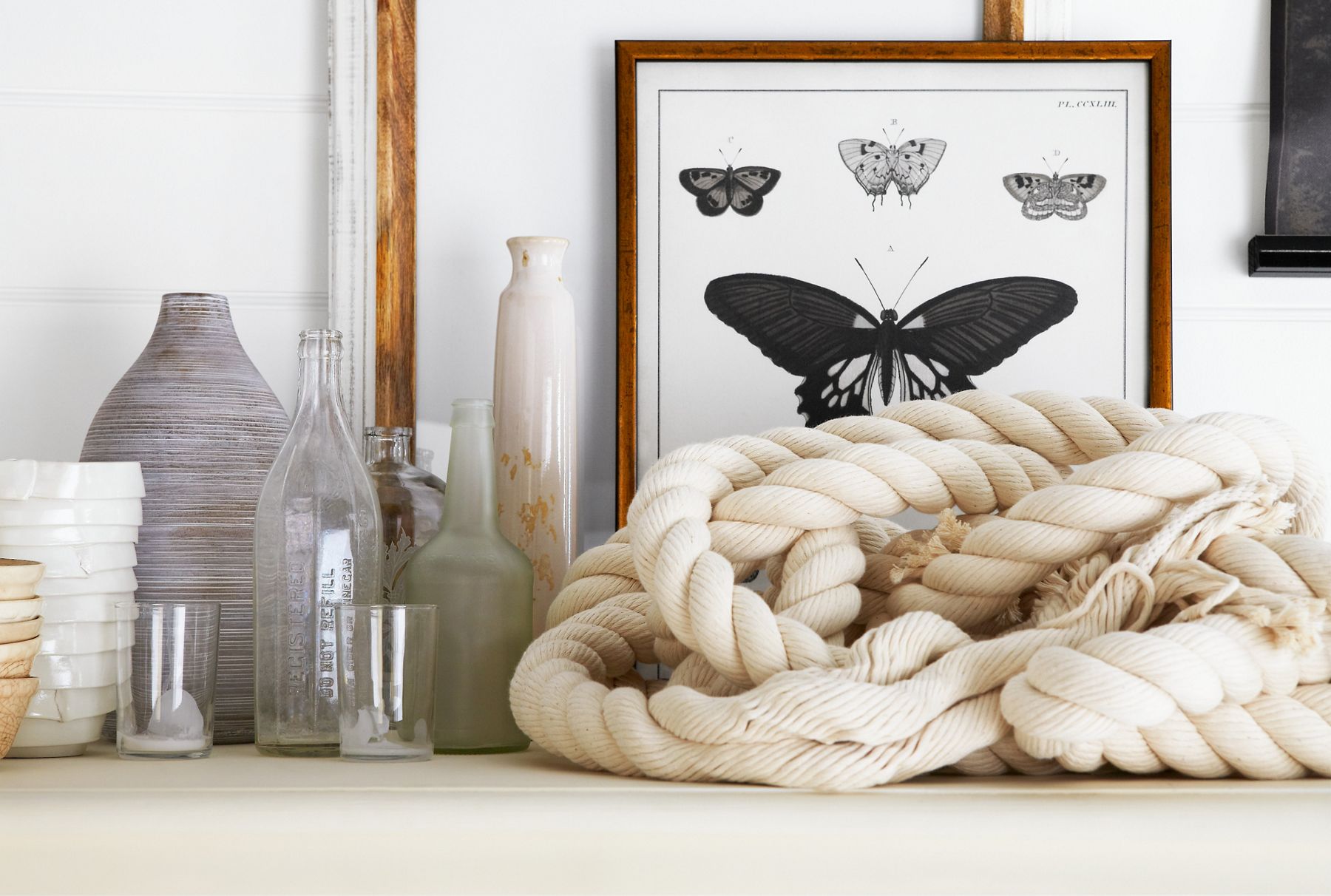 Detail of a display featuring glass and ceramic vases, framed black butterflies, and white rope for a coastal-inspired bath.