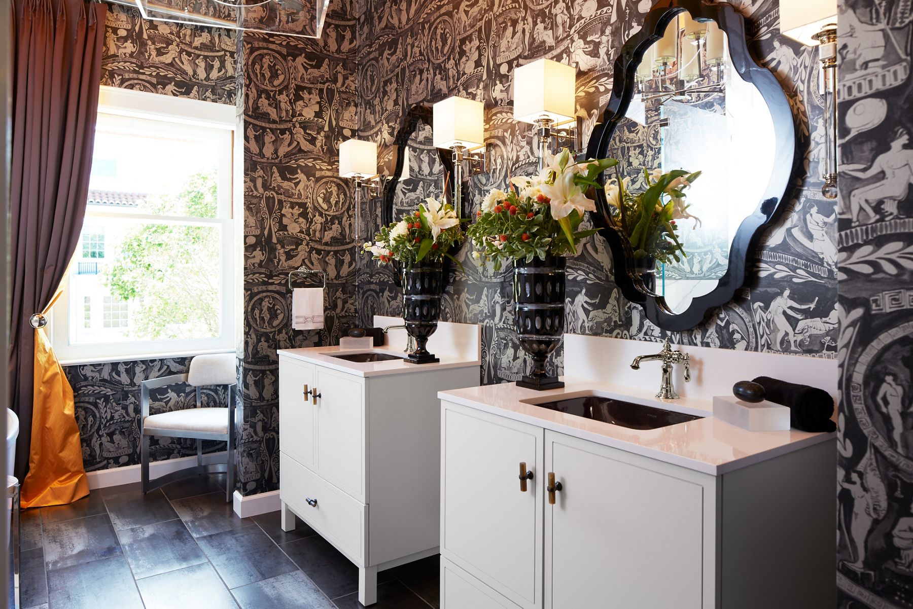 A polished chrome single-control Artifacts faucet on a white vanity top and with a black, scallop-framed mirror on a wall covered in wallpaper with imagery reminiscent of reliefs on ancient Roman pottery.