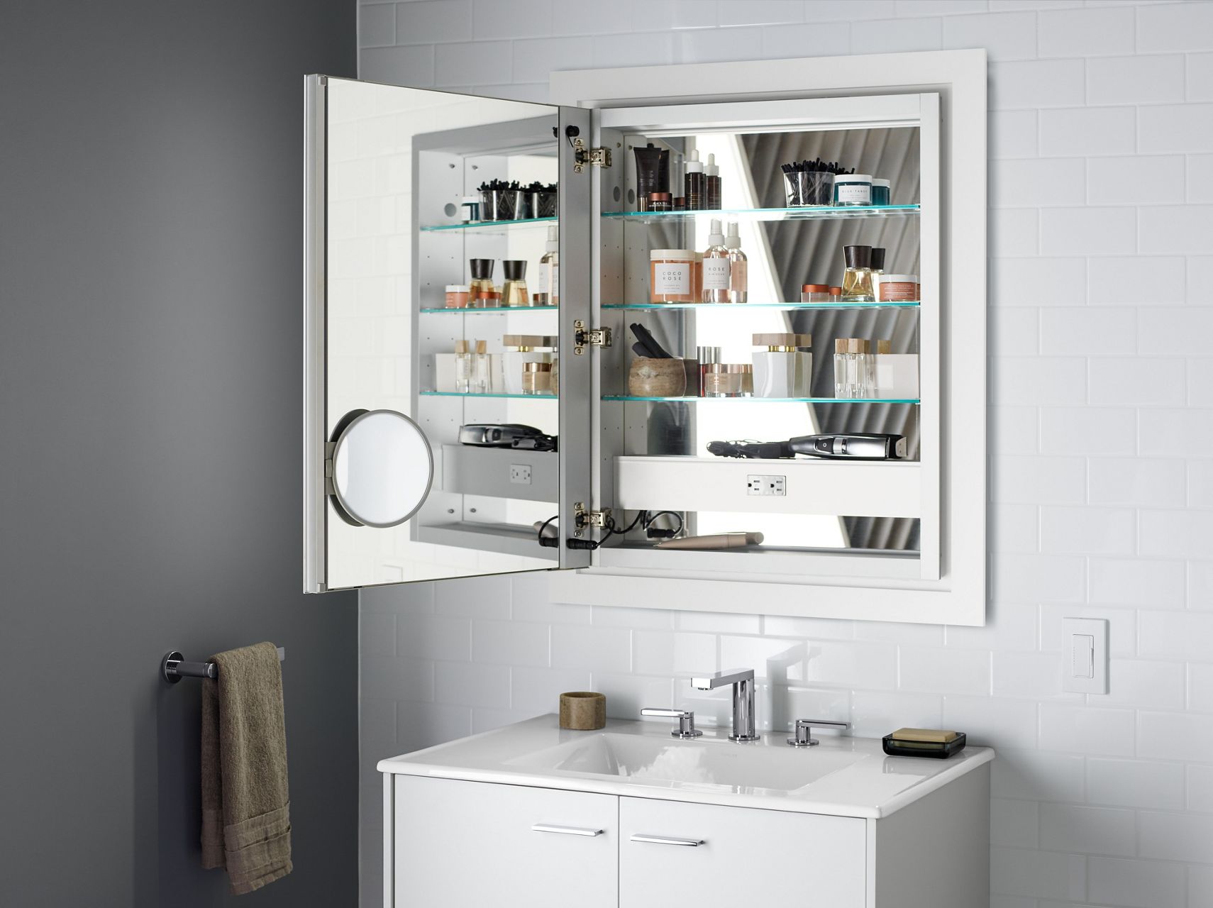 A KOHLER open medicine cabinet reveals available features, including a mirrored interior and an adjustable magnifying mirror.