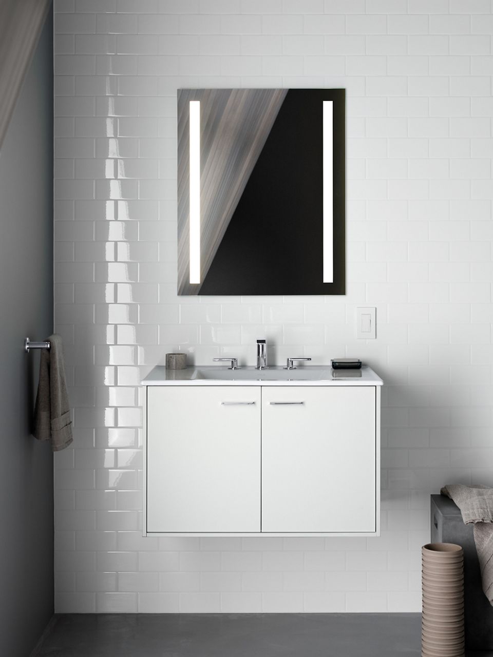 A lighted mirror hanging against a white tiled wall above a white vanity with a chrome faucet.