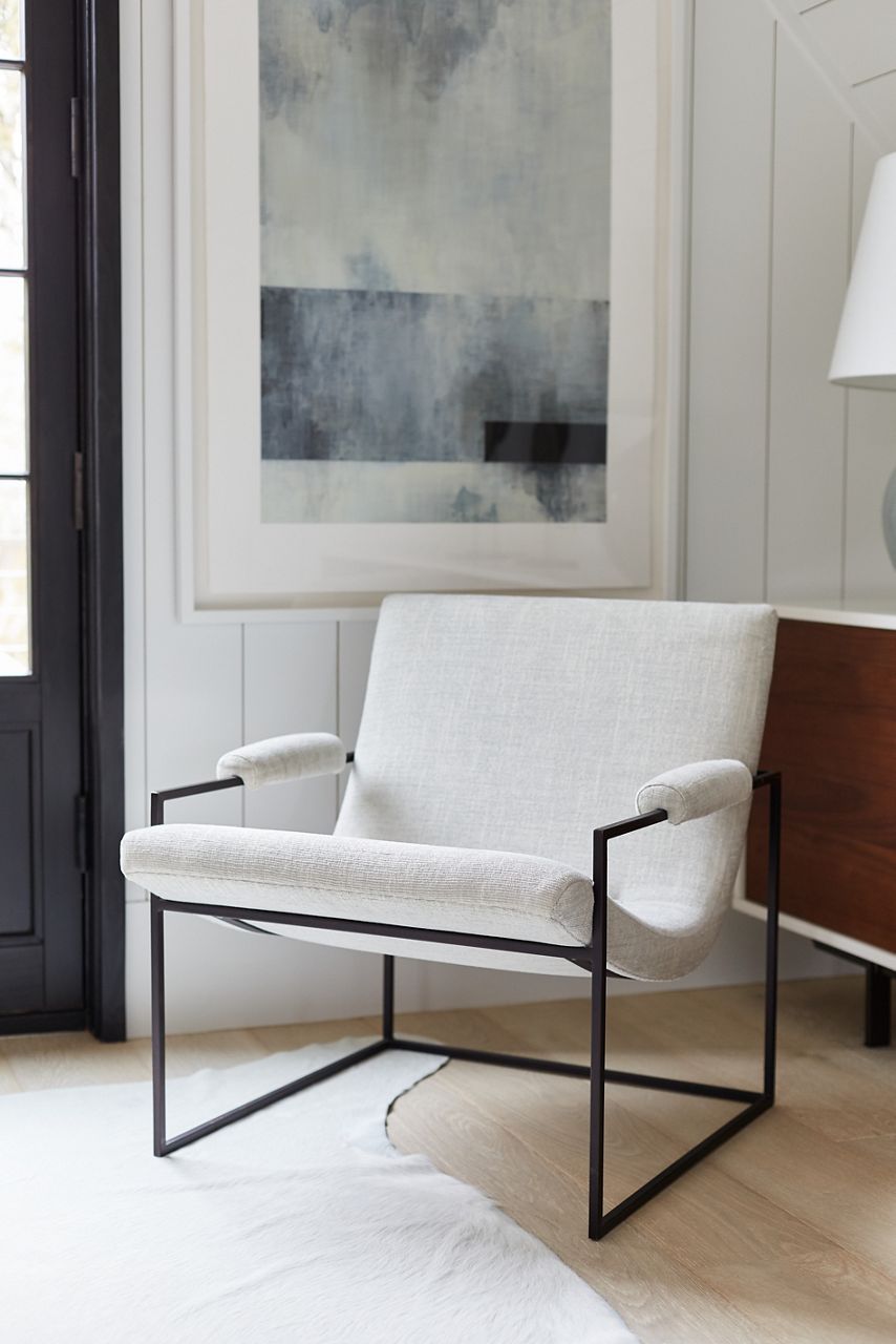 Un coin de la maison de Ray Booth à Provincetown, au Massachusetts, avec une chaise en cadre de métal noir avec un rembourrage blanc, un tapis blanc, des planchers en bois clair, des murs en planches blanches et une grande œuvre d’art abstraite dans des tons de blanc, de gris et de bleu.