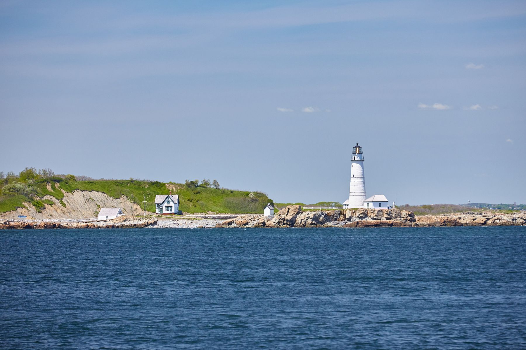 Une vue sur un phare blanc depuis l’eau, avec des dunes et une plage derrière elle.