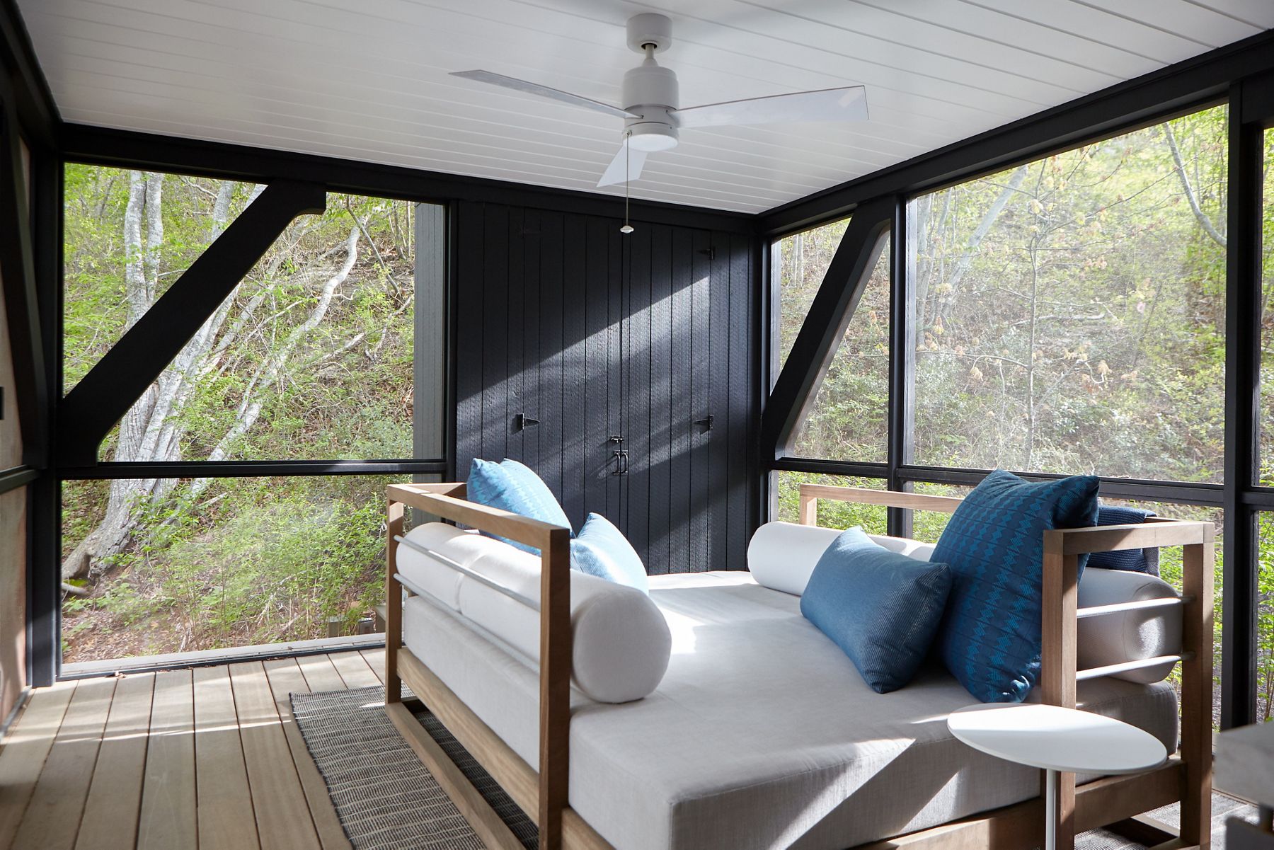 La lumière traverse les fenêtres du plancher au plafond dans une chambre avec de larges planchers en planches et un lit à cadre en bois avec des coussins blancs et bleus.