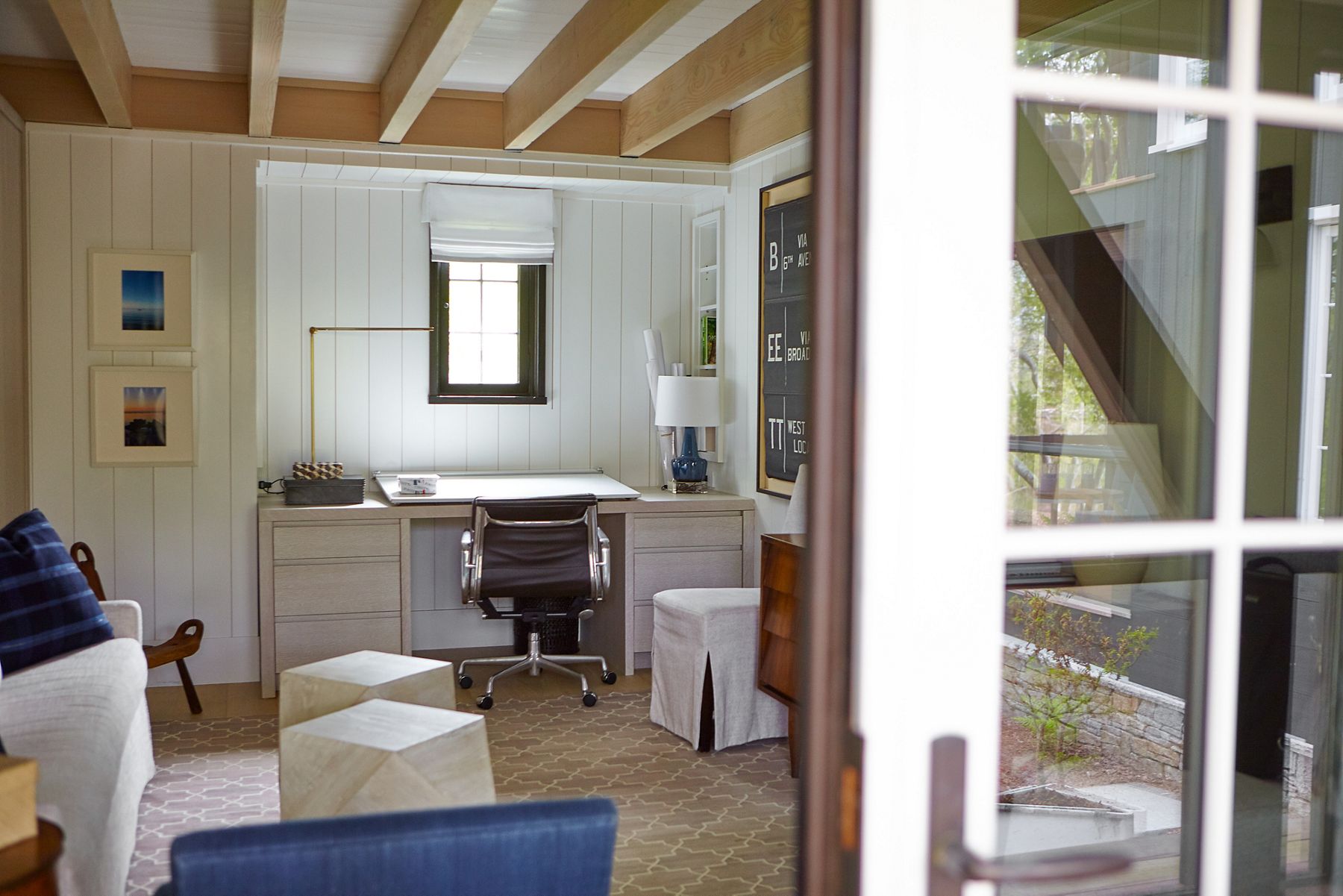 Un bureau à domicile, avec des murs à panneaux blancs, un bureau et une chaise de bureau sous une fenêtre avec un cadre noir, et un canapé blanc décontracté, une chaise bleue et des poufs blancs avec housse au lieu d’une table basse.