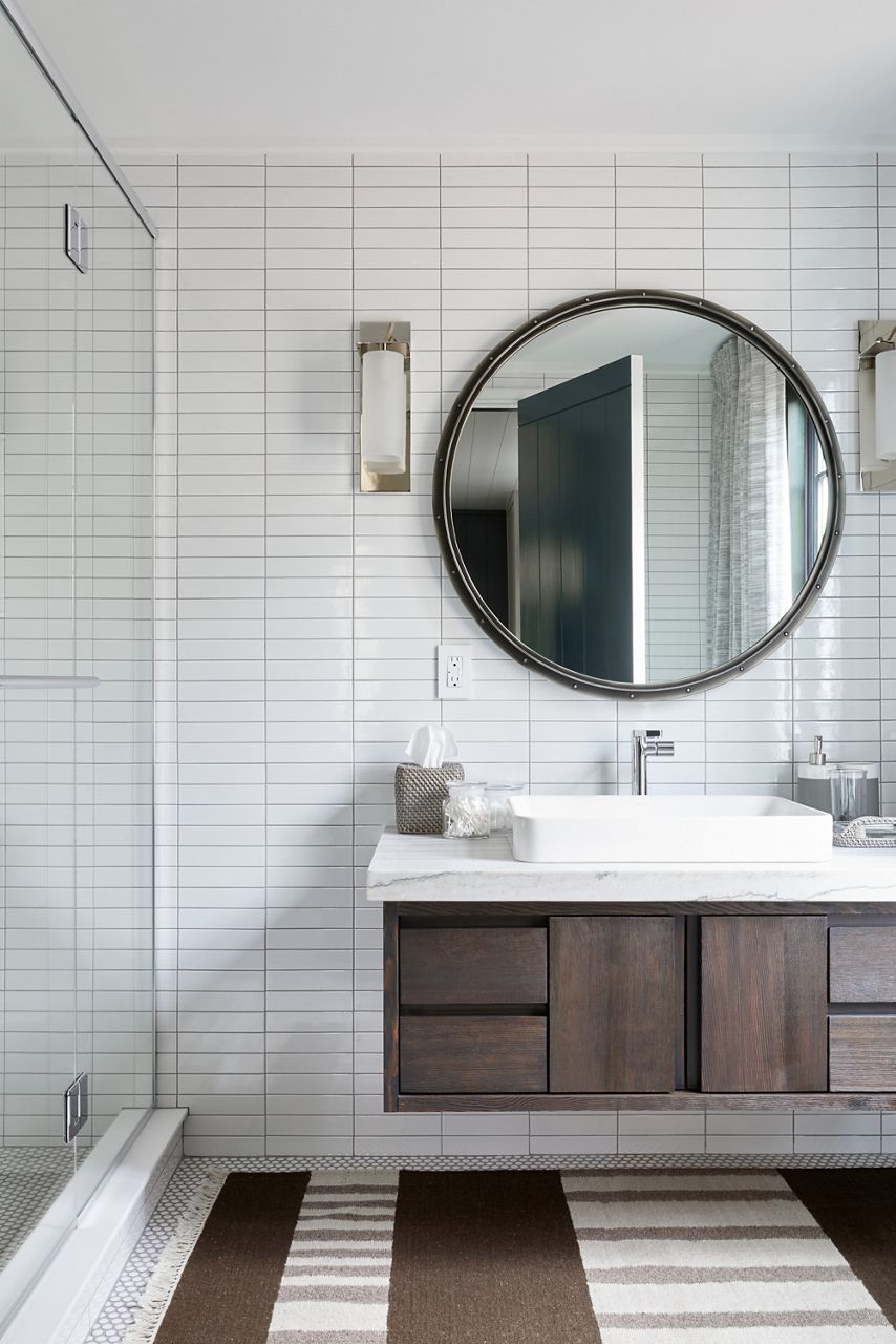 Une vue sur une salle de bain avec un tapis à motif neutre, un meuble-lavabo mural en bois foncé, un dessus de meuble-lavabo blanc avec lavabo en surface rectangulaire, un mur rectangulaire blanc et un grand miroir rond avec appliques murales cylindriques de chaque côté