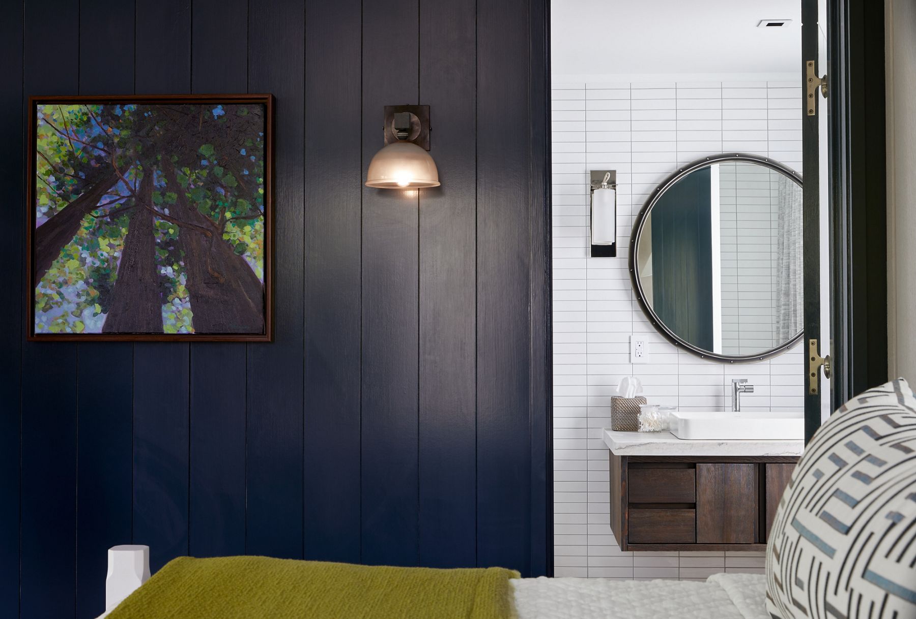 Une porte ouverte dans un mur à panneaux foncés révèle une salle de bain avec un miroir rond, des appliques murales, un meuble-lavabo suspendu à installation murale et un lavabo rectangulaire Vox de KOHLER.