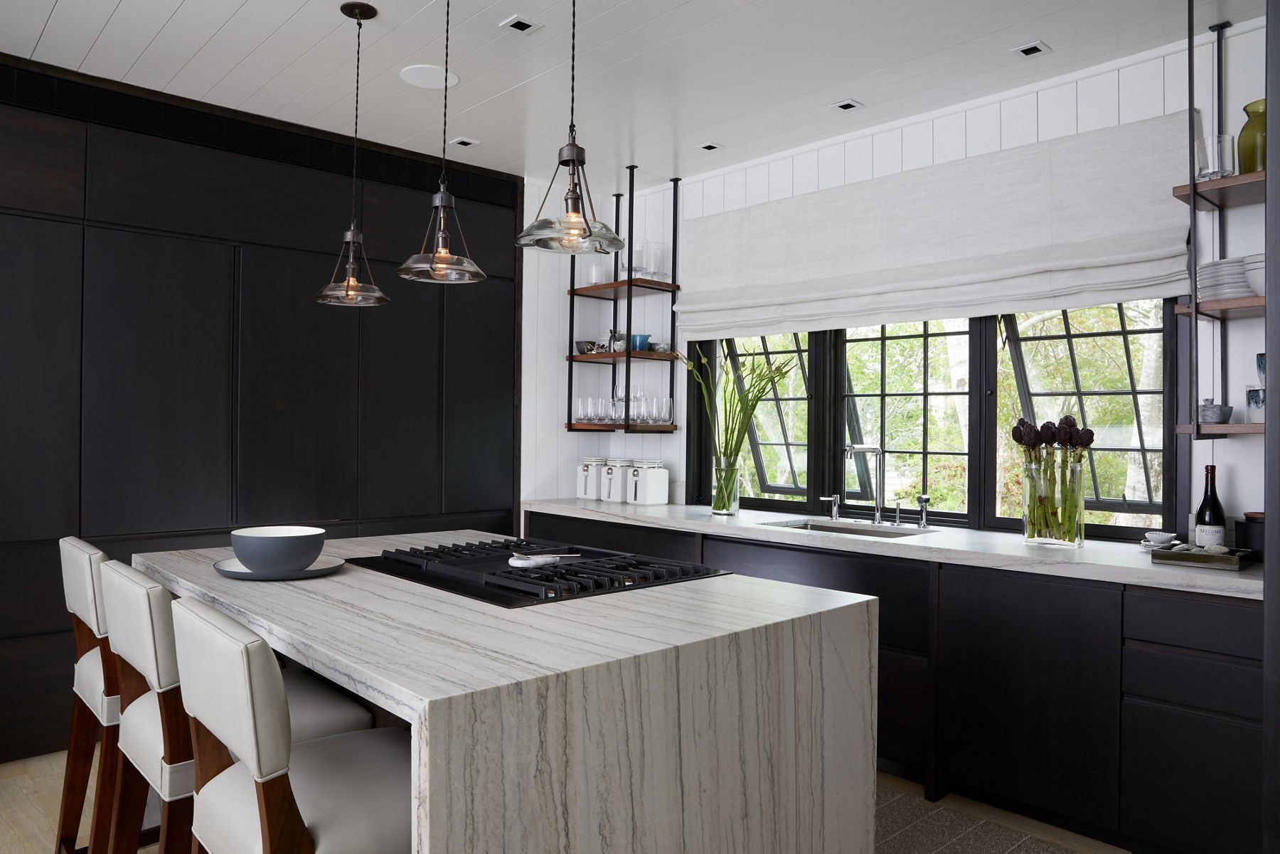 Vue sur une cuisine avec un mur d’armoires de plancher sombre à plafond, une grande île avec un comptoir en pierre en cascade et une table de cuisson montée sur l’île, et une rangée d’armoires inférieures sombres avec un comptoir blanc sous trois grandes fenêtres.