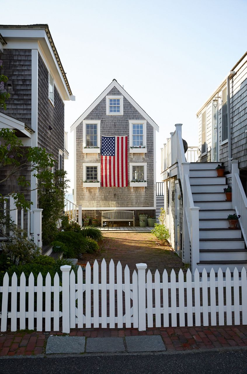 Une maison traditionnelle en cèdre Cape Cod avec un grand drapeau américain suspendu entre ses fenêtres avant et une clôture blanche en forme de piquet devant elle.