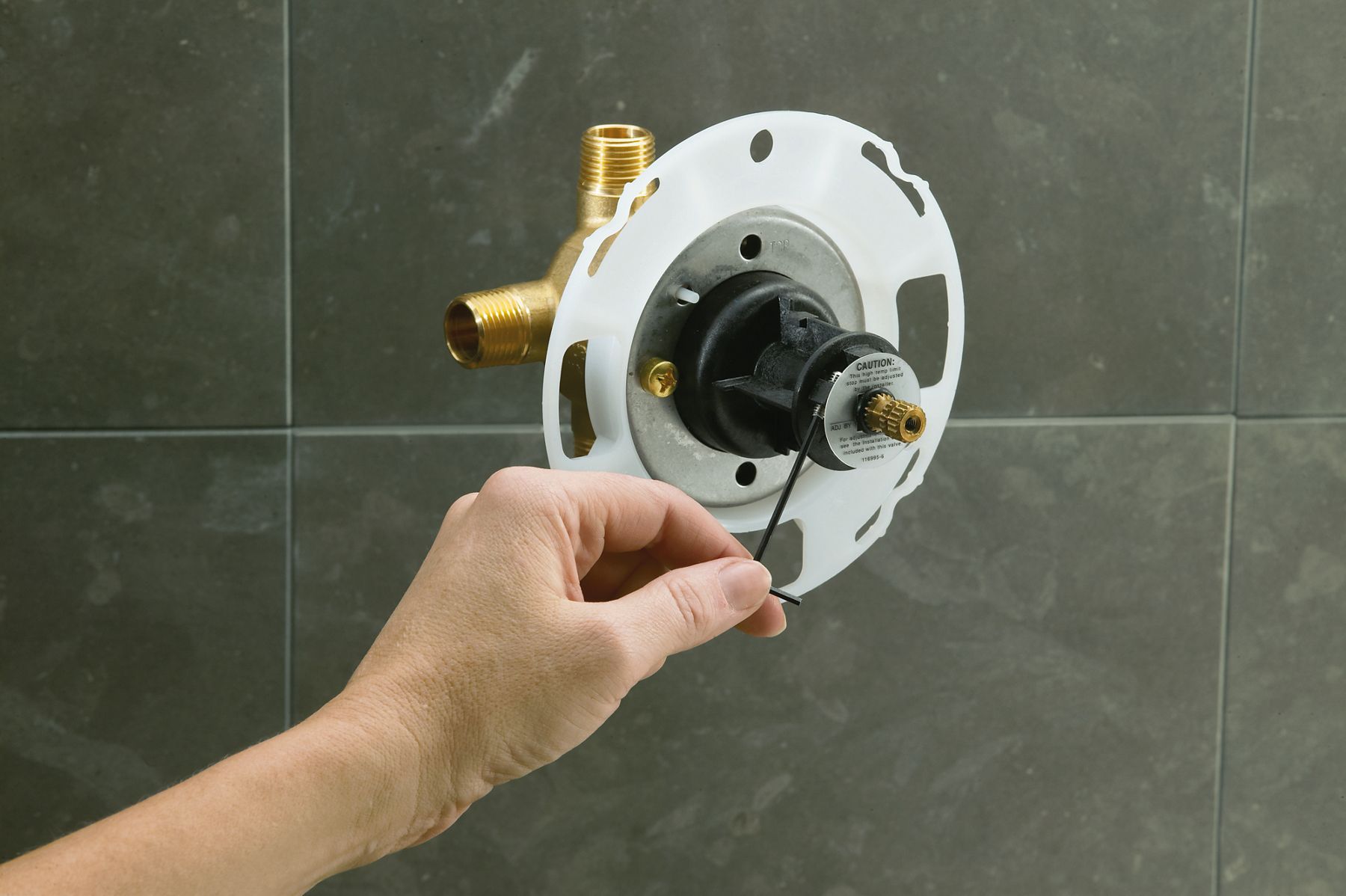 Hand adjusting a shower valve rough-in installation with white plaster guard mounted against dark gray tiled shower wall.