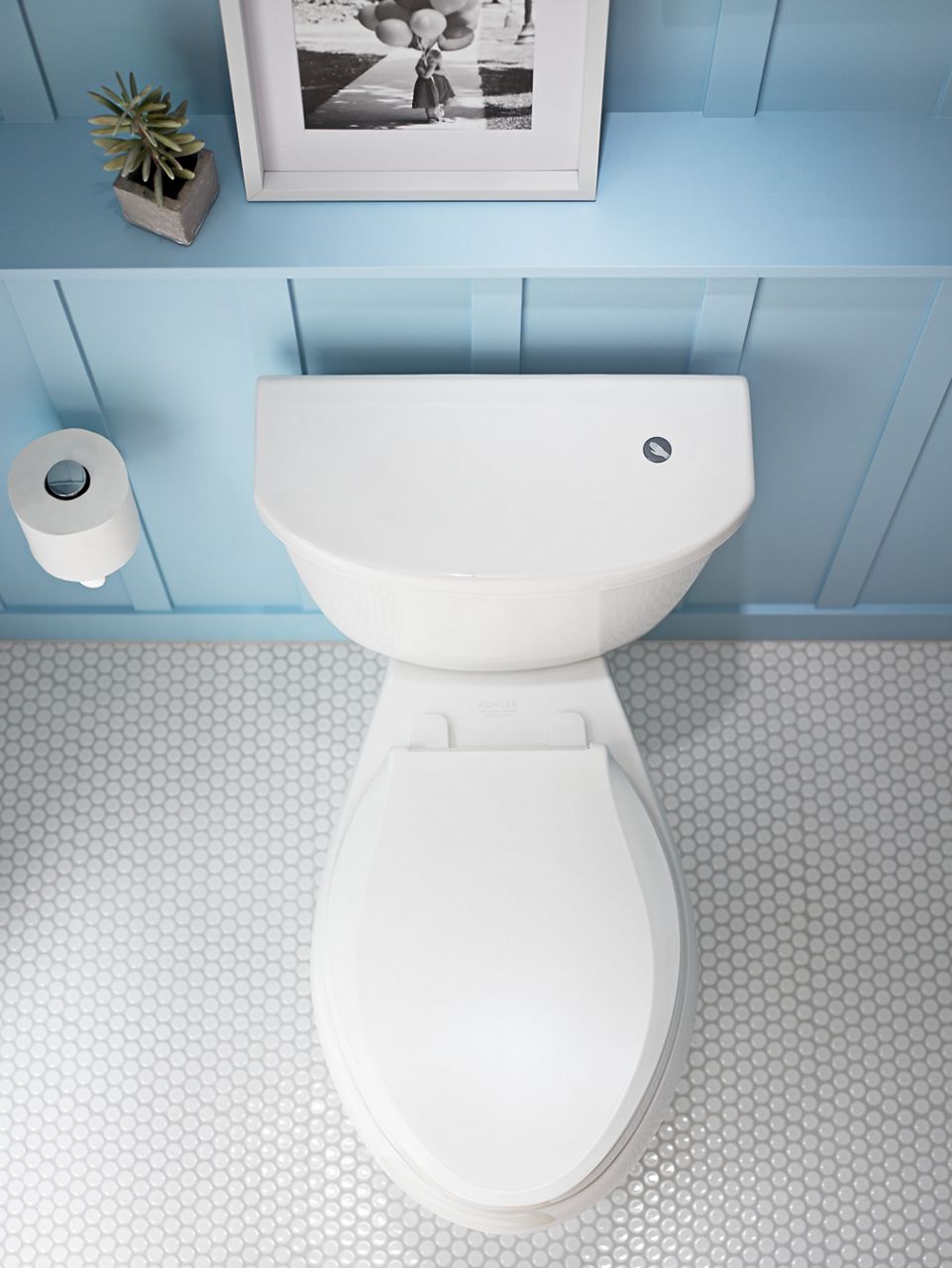 A top-down view of a white toilet and seat on a floor of small white hexagonal tiles and dark grout.