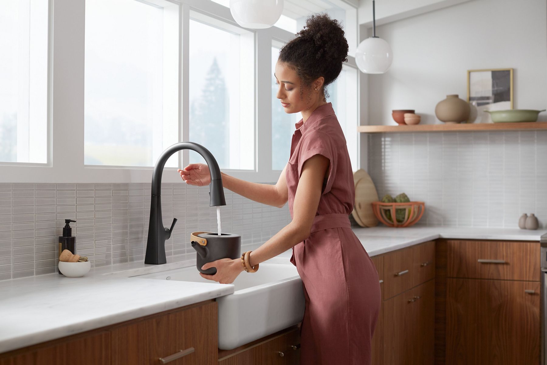 A woman waving her hand under a touchless faucet as it pours water into a tea kettle.