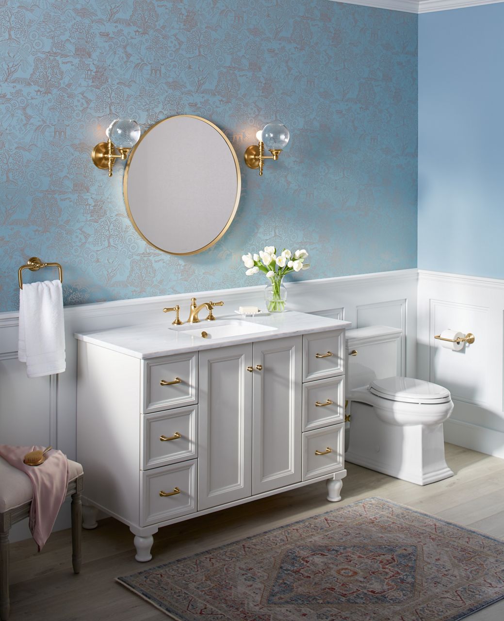 A tidy, well-organized bathroom with large white KOHLER vanity, widespread lever handle faucets, towel ring, and round mirror flanked by two sconces.