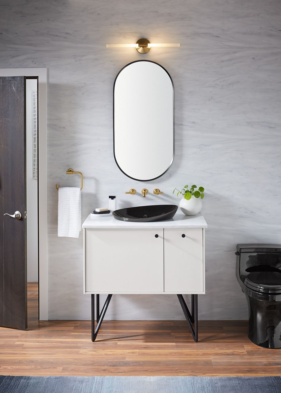An oval-shaped mirror with rounded top and bottom and straight sides against a marbled gray wall, with a two-light sconce above it and a brushed brass wall-mount faucet and vessel sink below.