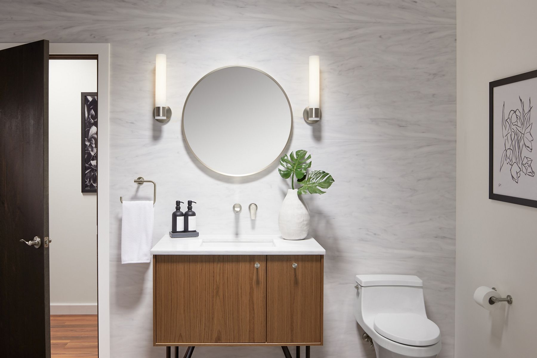 A brushed nickel-framed circular mirror with two sconce lights on either side on a white wall with a brushed nickel wall-mount faucet above a wood vanity with white counter and sink.