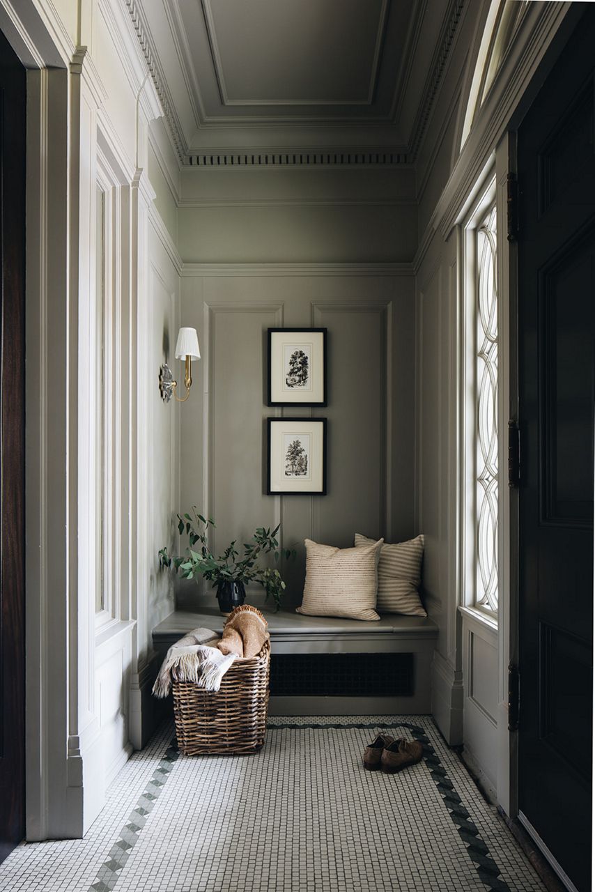Light streams through a window in sage green paneled nook, with bench, penny tile mosaic floor, framed art, and a basket of blankets.