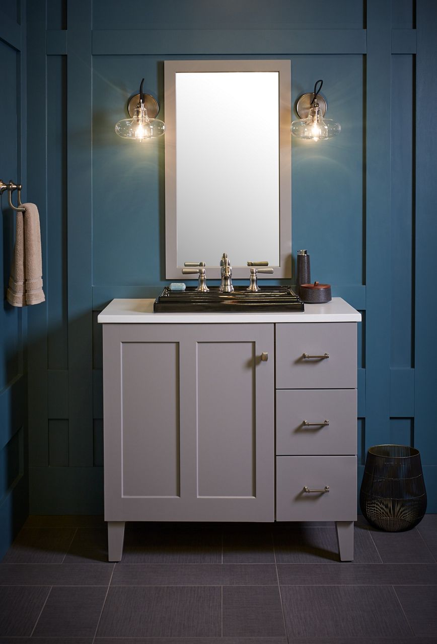 A bathroom styling space with a gray vanity with three drawers on the right and a large door on the left, a white counter top, matching rectangular mirror and sconce lights on a deep teal wall. 