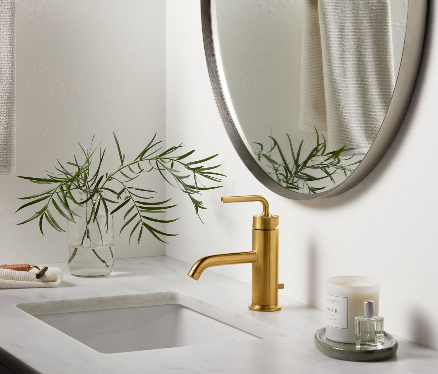 A white bathroom vanity top with accessory tray, vase with branches, and a KOHLER Purist single lever handle faucet in Vibrant Brushed Moderne Brass.