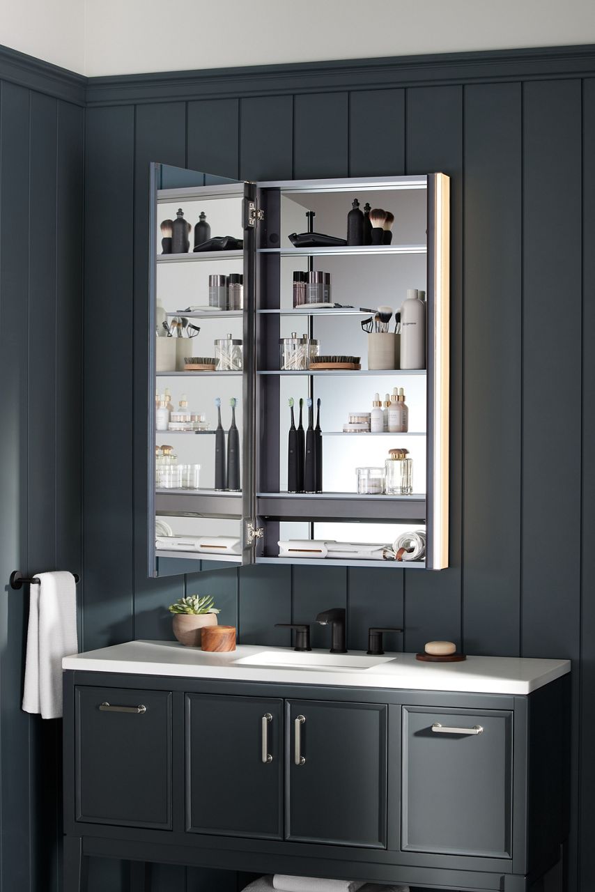 An opened Maxstow medicine cabinet, revealing staggered shelving and dual interior mirrors, in a bathroom with dark gray walls and a dark gray vanity with a white top and black faucet.
