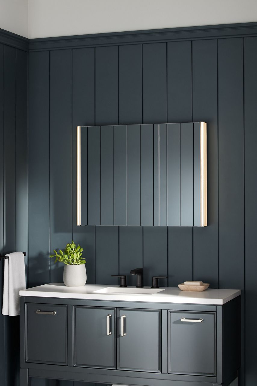 A KOHLER Verdera lighted medicine cabinet shines in a moody bathroom with black paneled walls, dark gray vanity, and white vanity top.