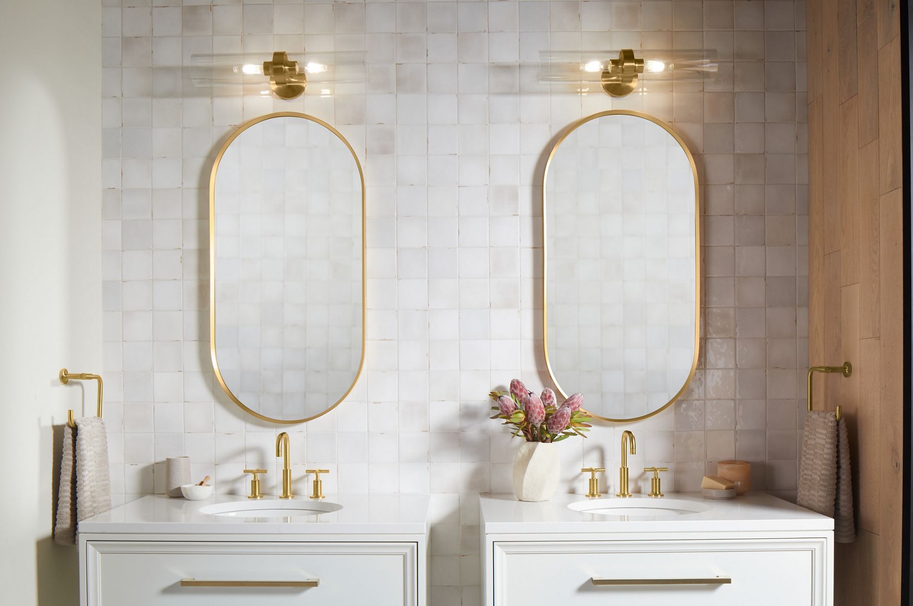 A bathroom with square white tiled wall, two white KOHLER vanities with widespread lever handle faucet in Vibrant Brushed Brass Moderne, and two capsule mirrors above them.