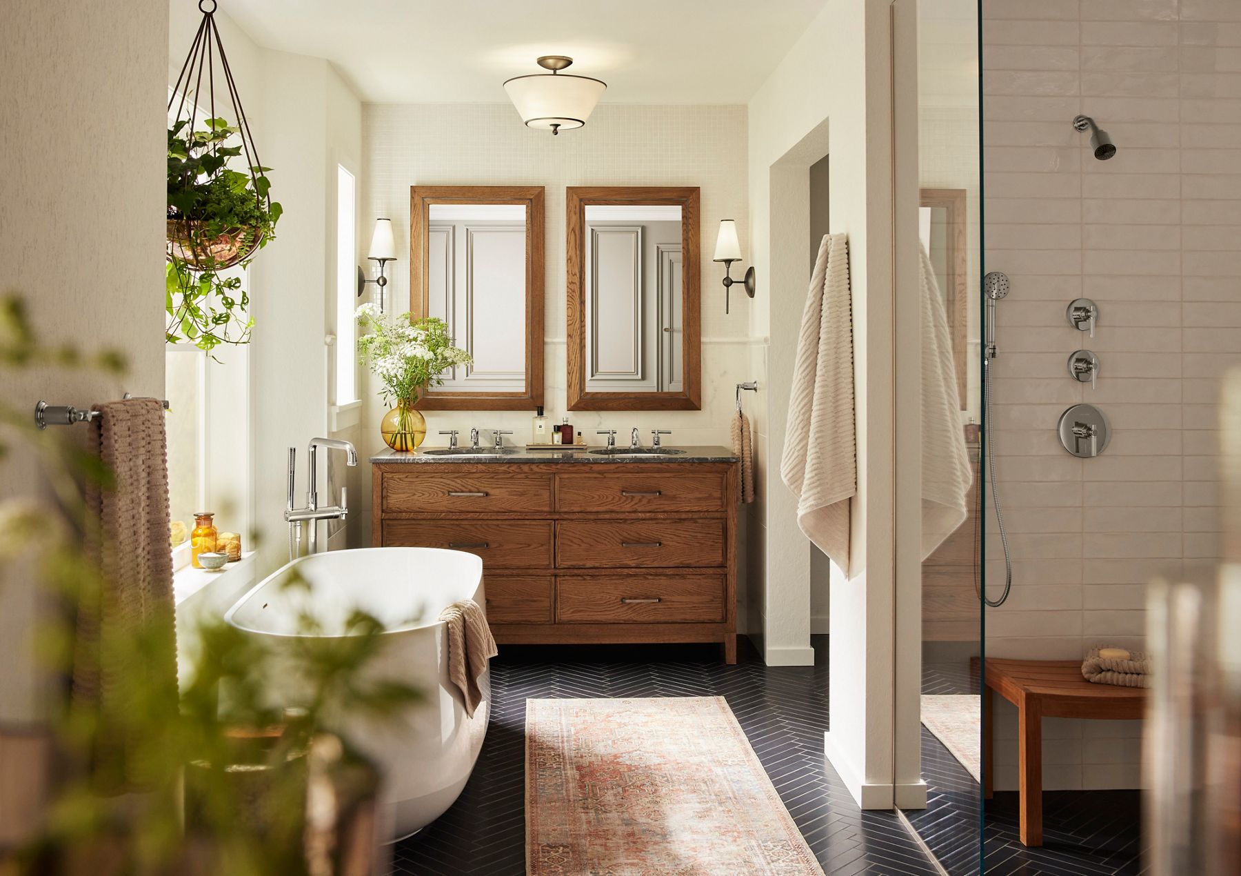 Elegant bathroom interior featuring a freestanding white bathtub with a towel draped over the edge, positioned next to a wooden double vanity with two large framed mirrors above it. The room is softly lit by wall sconces on either side of the mirrors and a central ceiling light. A potted plant in a golden vase and fresh flowers add a touch of greenery, while a neutral-toned rug lies on the dark herringbone-patterned floor. Light, cream-colored walls and ample natural light create a serene and cozy atmosphere.