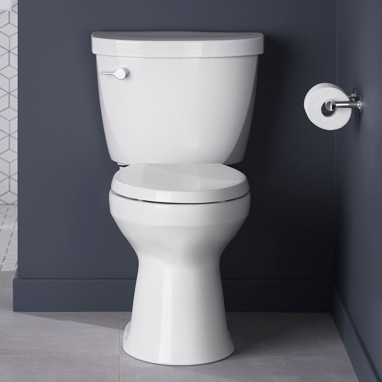 Vue de face d’une toilette Kohler blanche à côté d’un rouleau de papier hygiénique devant un mur gris foncé