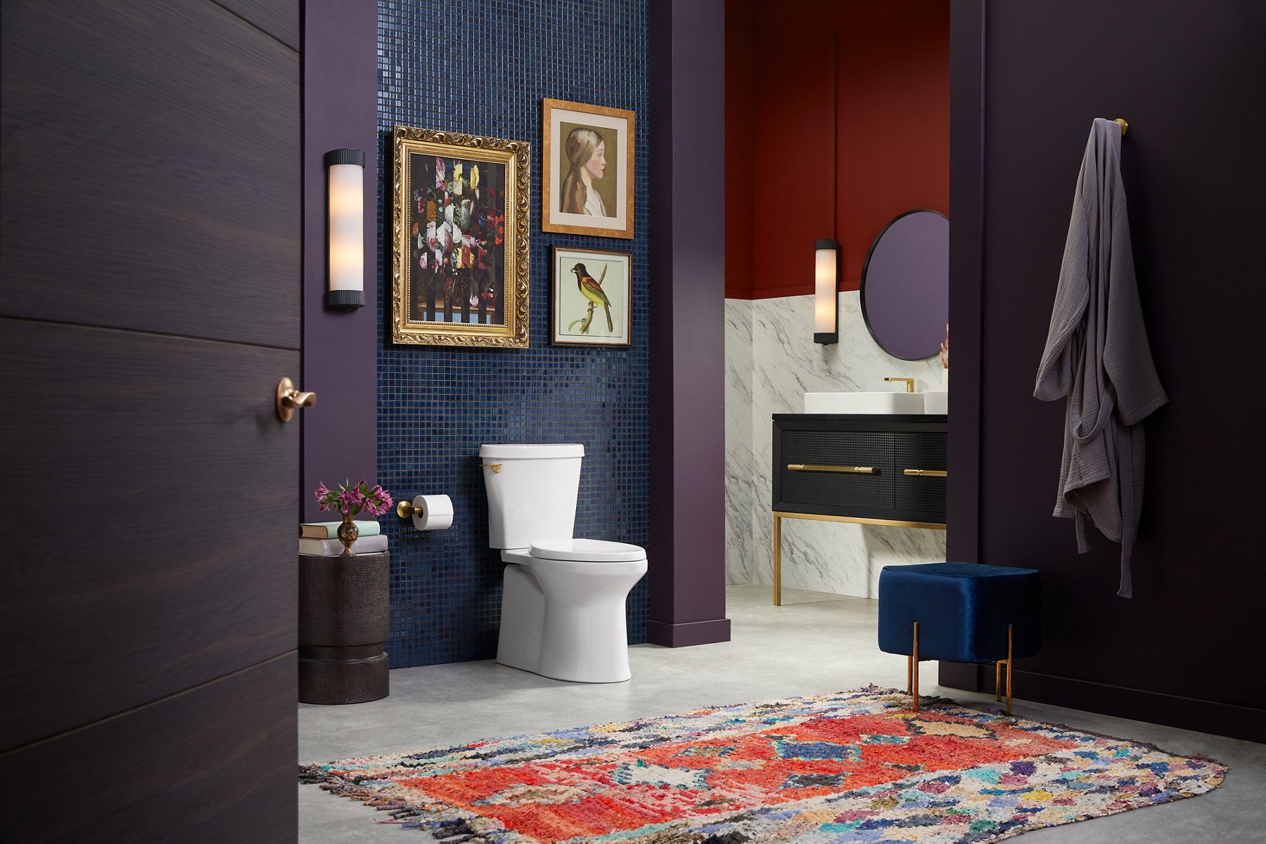 A transitional two-piece toilet wit ha skirted trapway and brass-colored flush actuator set against a shimmery blue tile wall.
