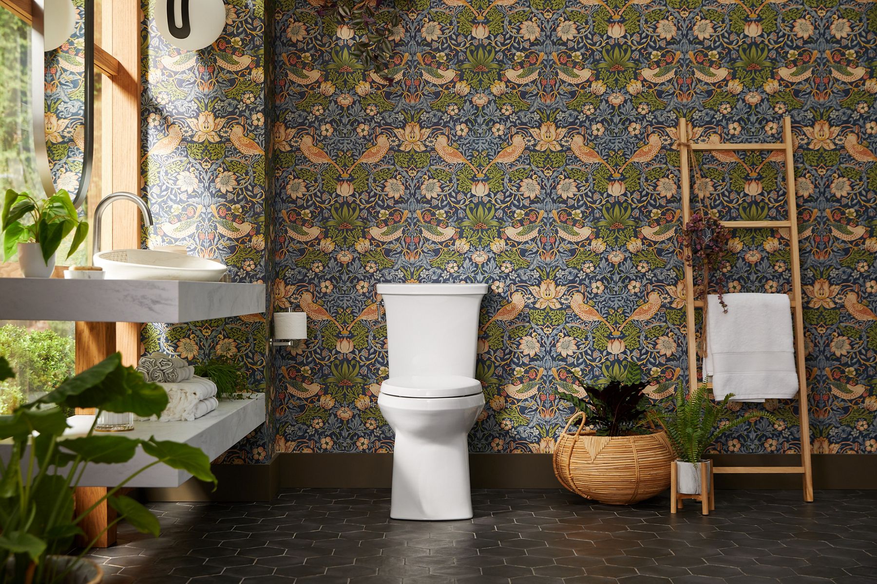 A cheerful, eclectic bathroom with large windows, patterned wallpaper, towel ladder, and a white KOHLER two-piece toilet.