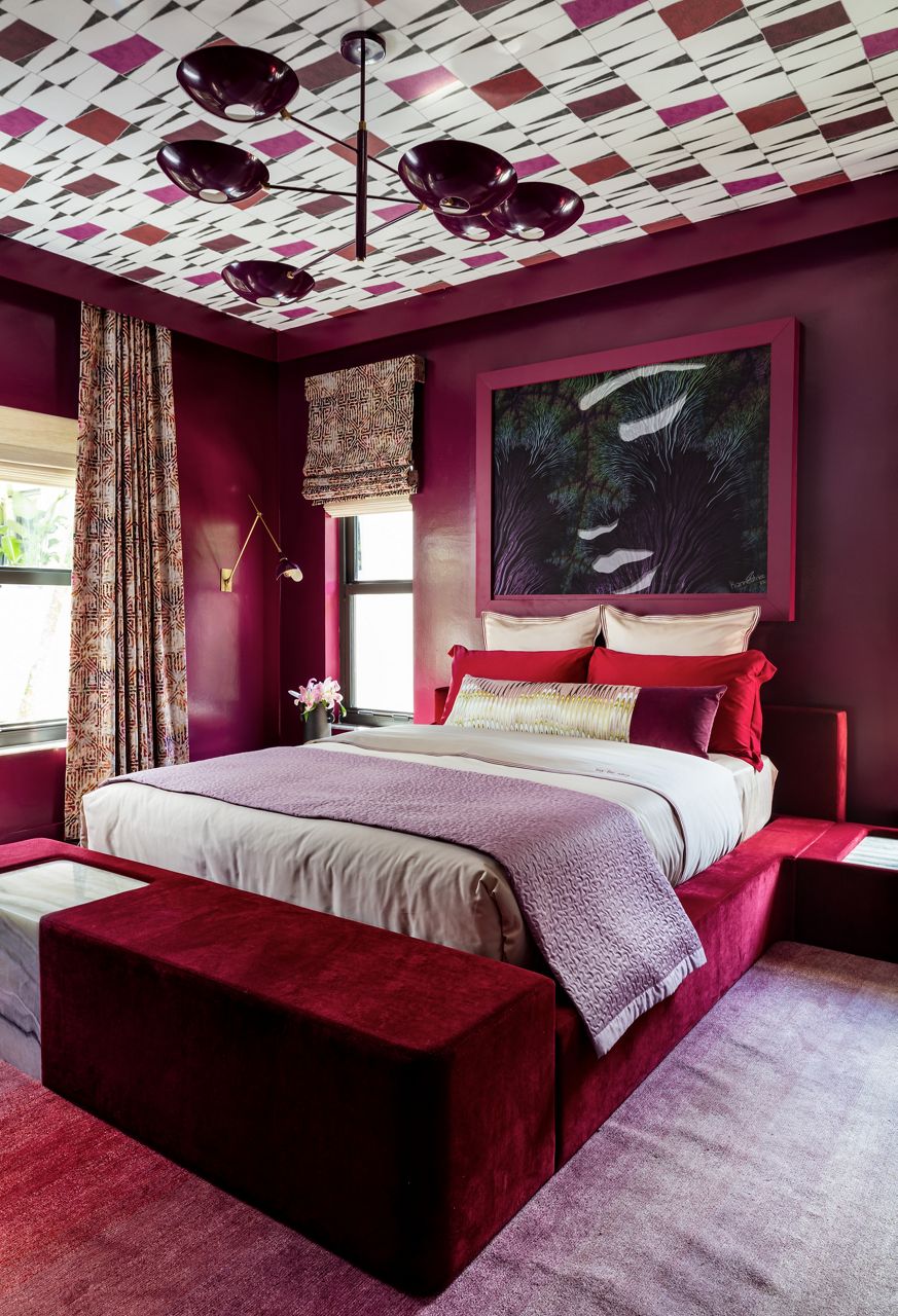 A bedroom in Curated Maximalist style, with burgundy walls, red platform bed with white cover and purple throw, chandelier, and red patterned wallpaper on the ceiling.