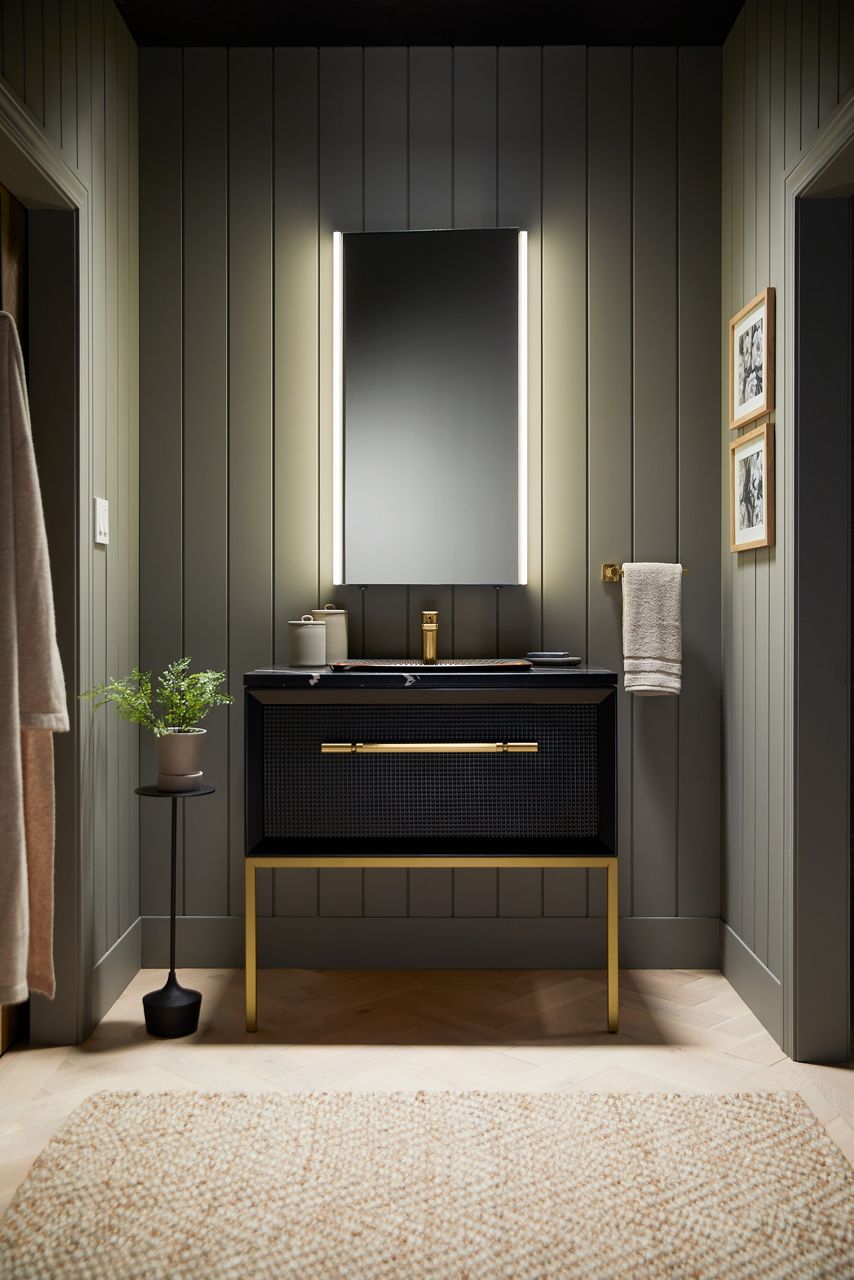 A bathroom with sage green paneled walls features a KOHLER lighted mirror, single lever handle faucet, and single drawer vanity in dark wood on metal furniture legs.