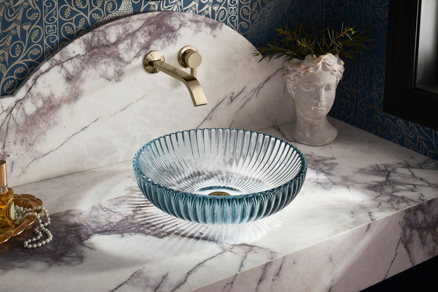 Un robinet mural au fini French Gold au-dessus d’un lavabo en surface en verre bleu cannelé sur un comptoir marbré violet et blanc.